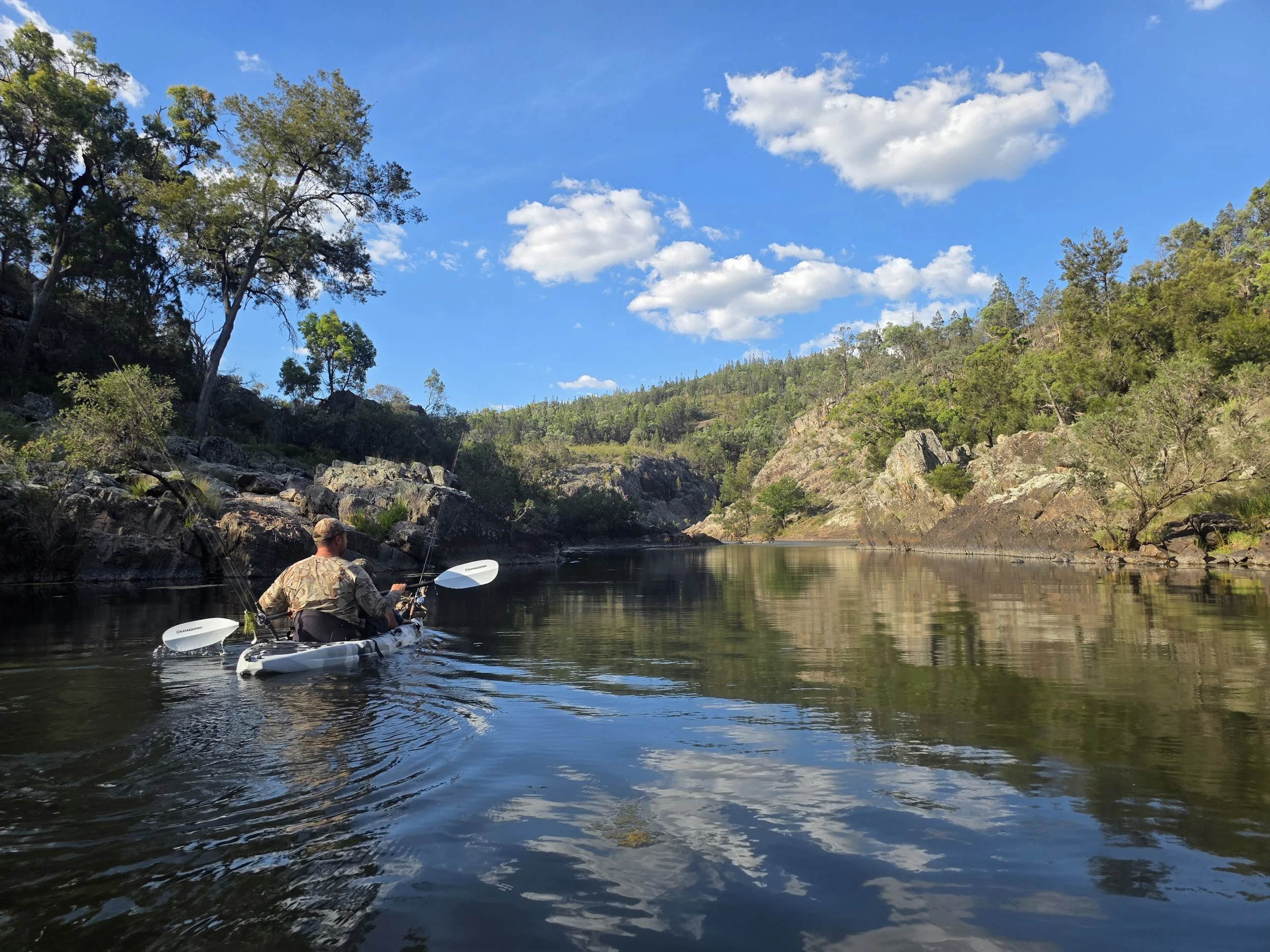 A person kayaking on a river, surrounded by rocky hills and green trees, under a bright blue sky with scattered clouds.