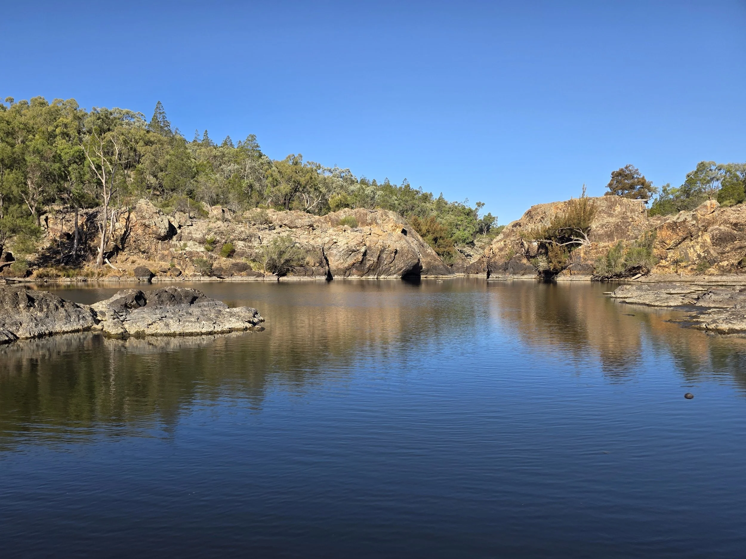 A calm river with large rocks along the shoreline, surrounded by green trees and a clear blue sky.