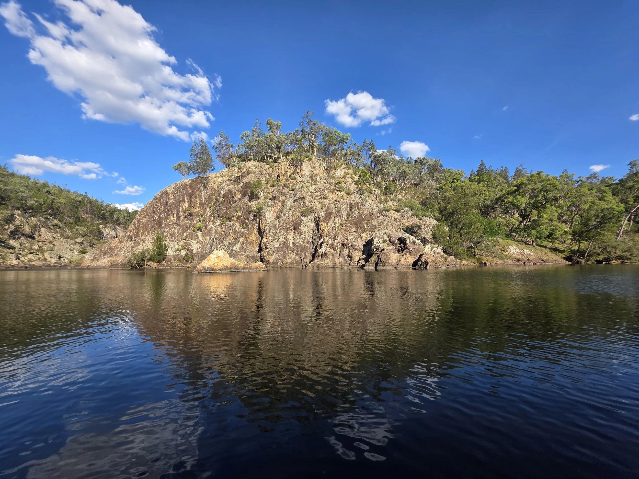 A rocky hill with green trees on top is reflected in calm water under a blue sky with scattered clouds.