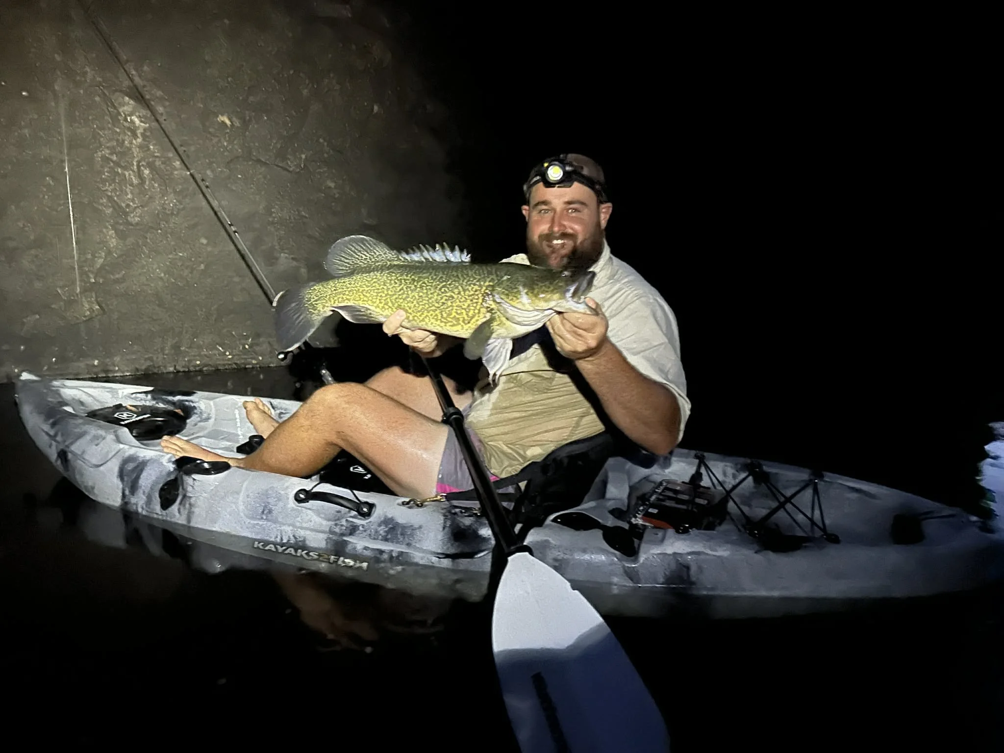 A man sitting in a kayak at night, holding a large fish with a fishing rod beside him, smiling at the camera.
