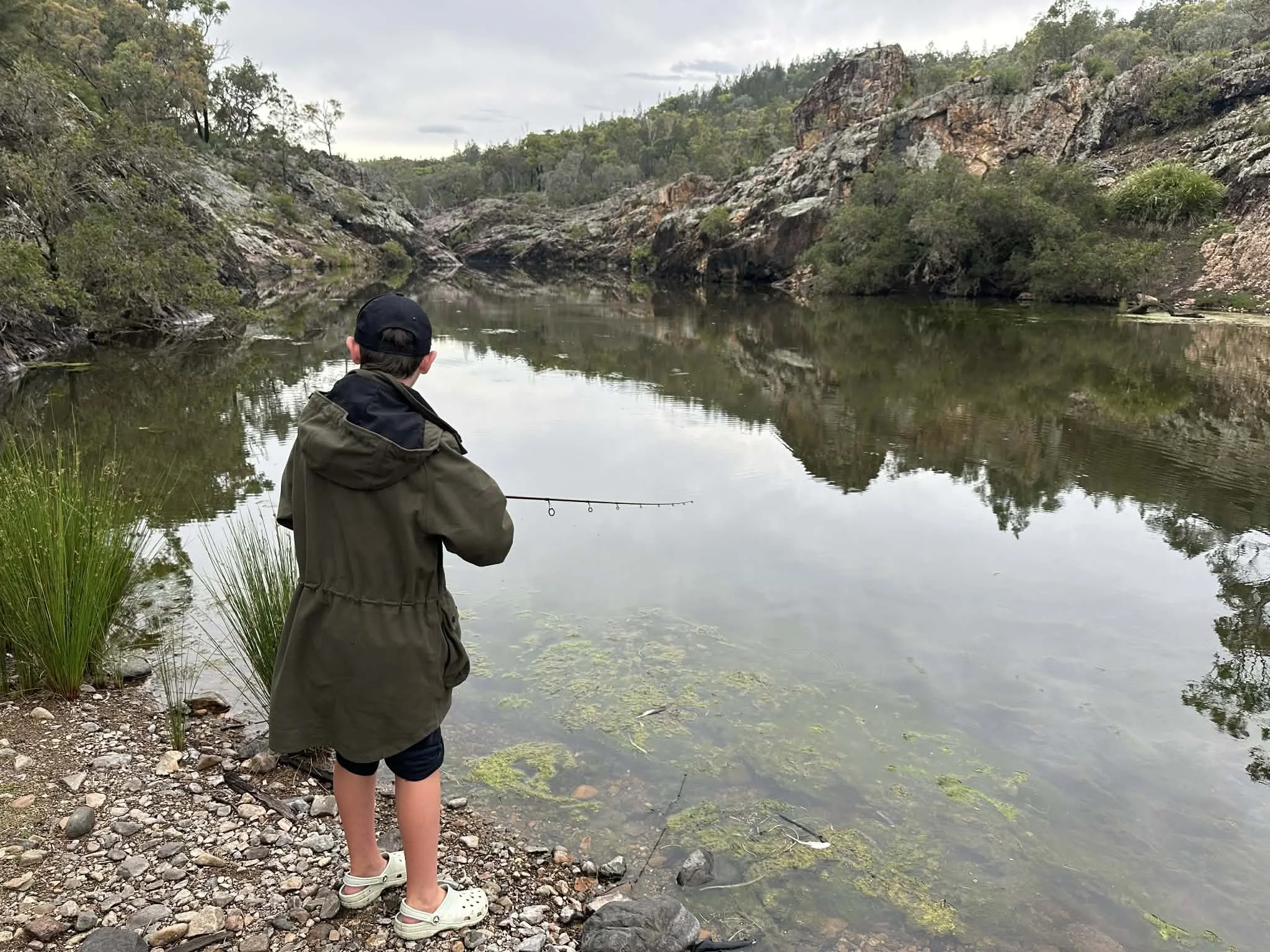 A young boy fishing in a river surrounded by rocky cliffs and green vegetation.
