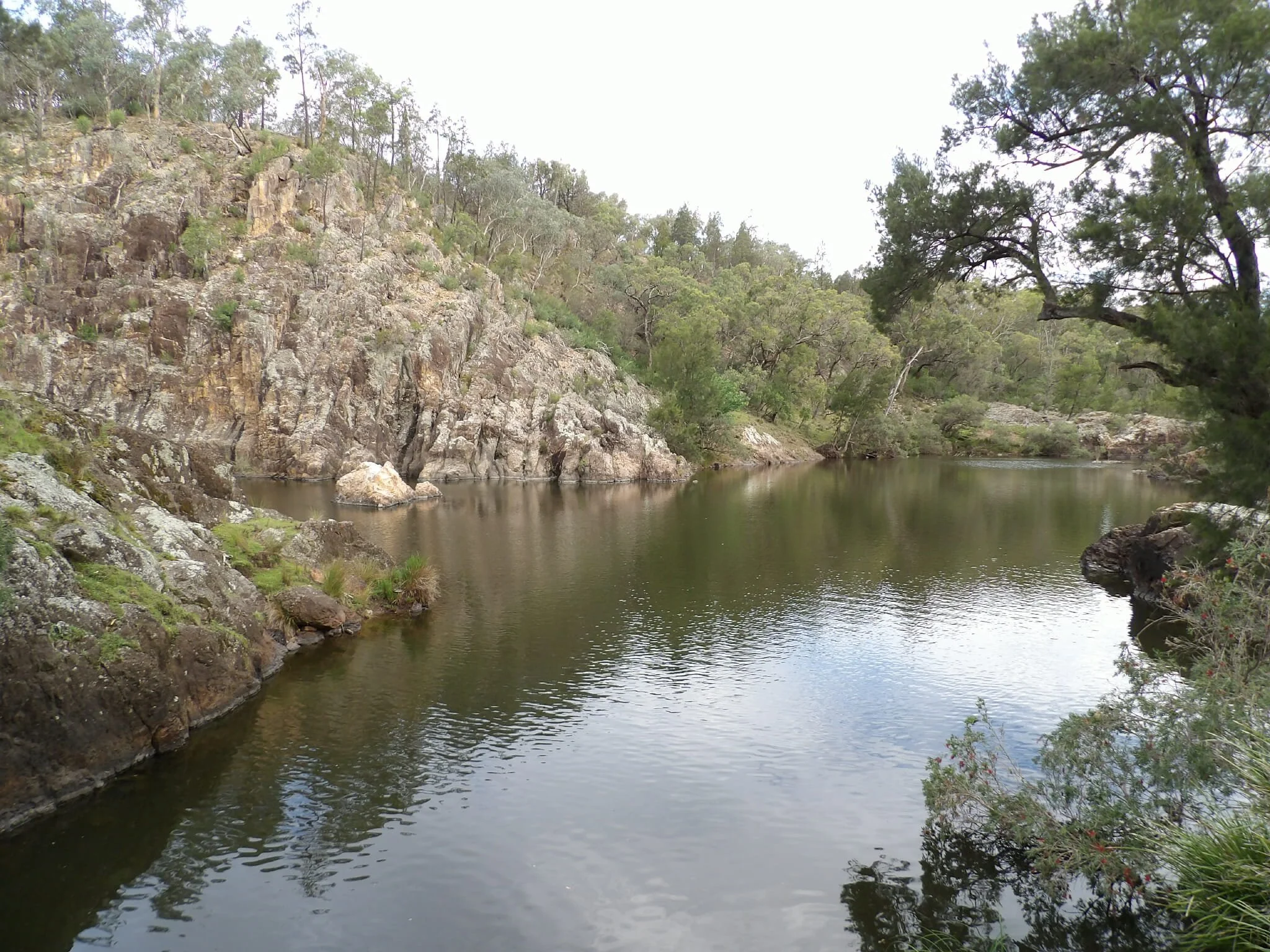 A peaceful river flowing through a rocky and wooded landscape with trees on both sides and a cloudy sky above.