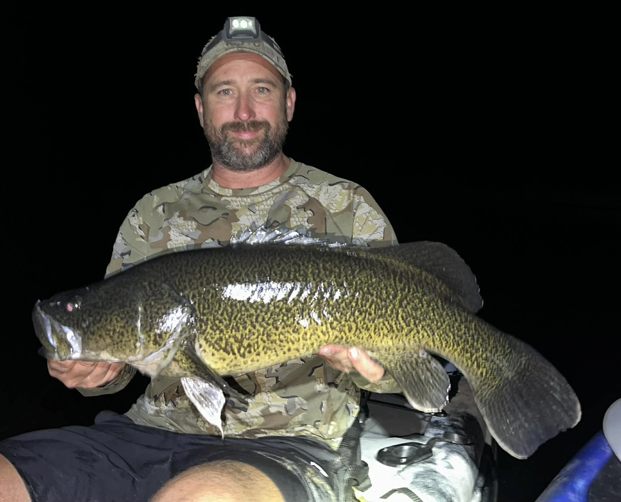 A man in camouflage clothing and a headlamp holding a large fish at night.
