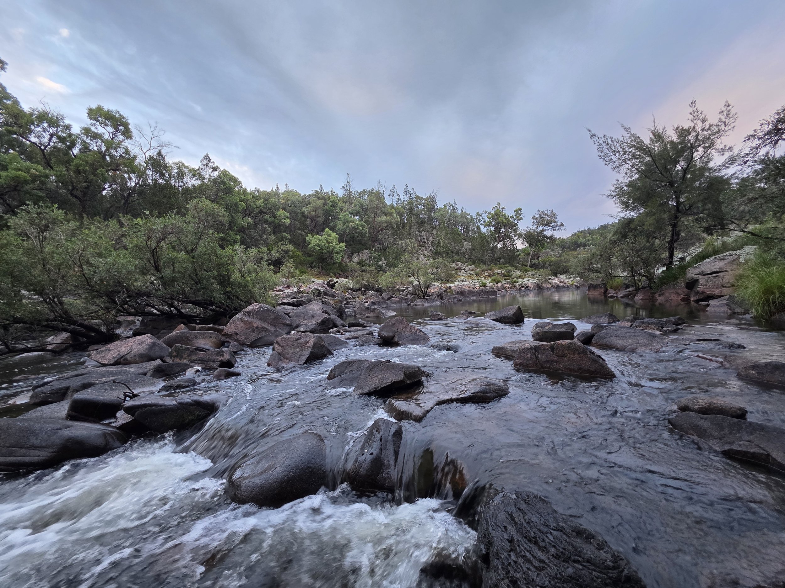 A rocky river flowing through a lush green forest under a sky with some clouds.