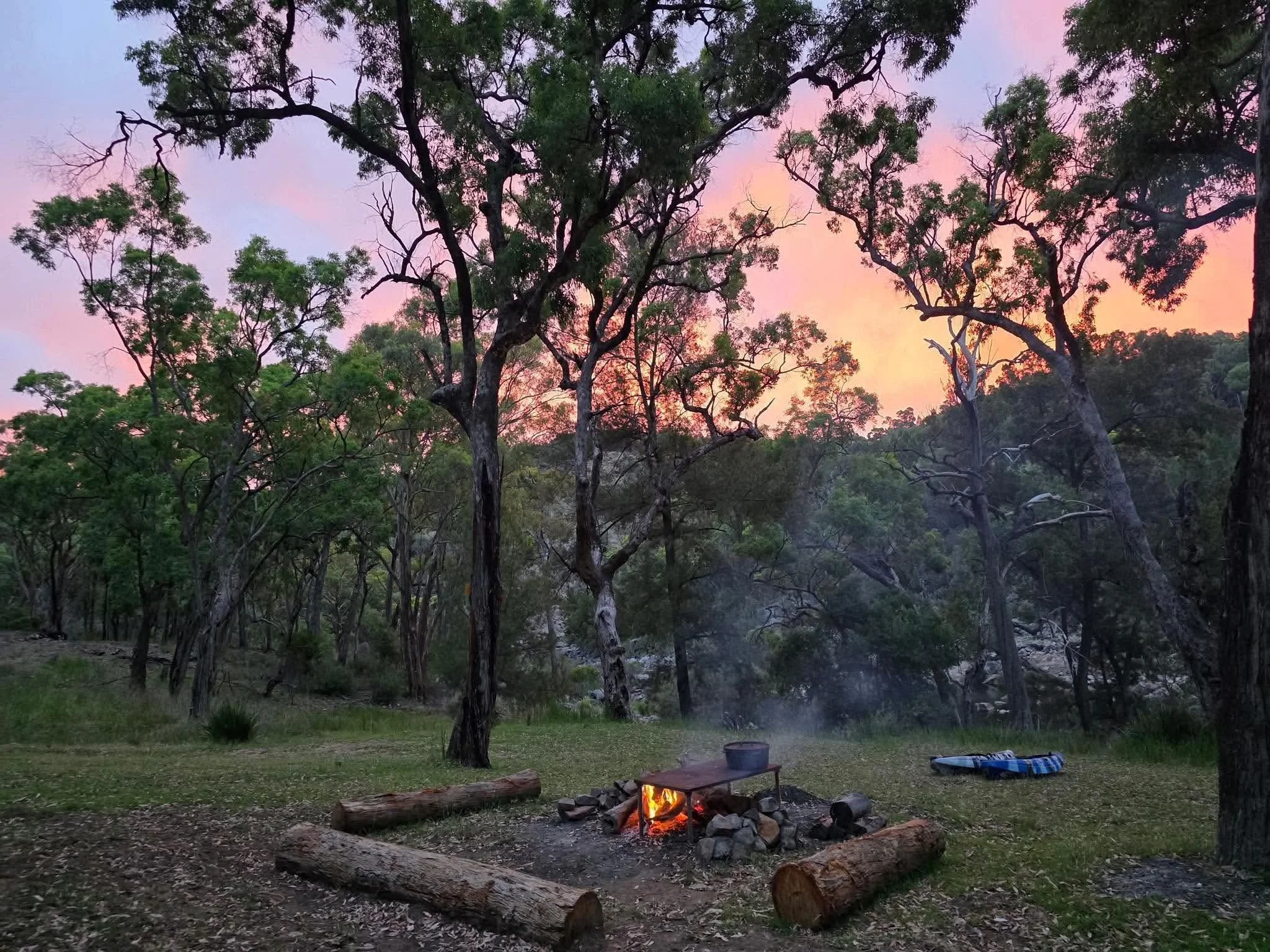A campfire at the Basecamp at the Severn River Outpost with logs and stones on the bank of the Severn River in New South Wales at dusk, with trees and a colorful sky in the background.