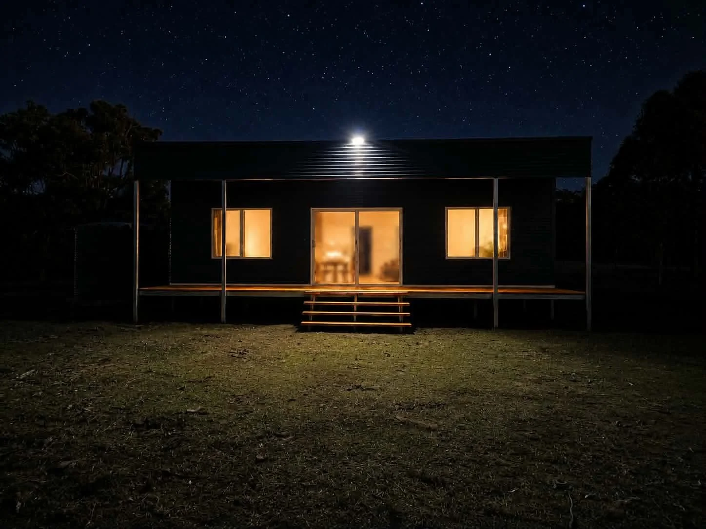 A modern house with large windows and a porch, illuminated from inside, under a starry night sky with a bright moon.