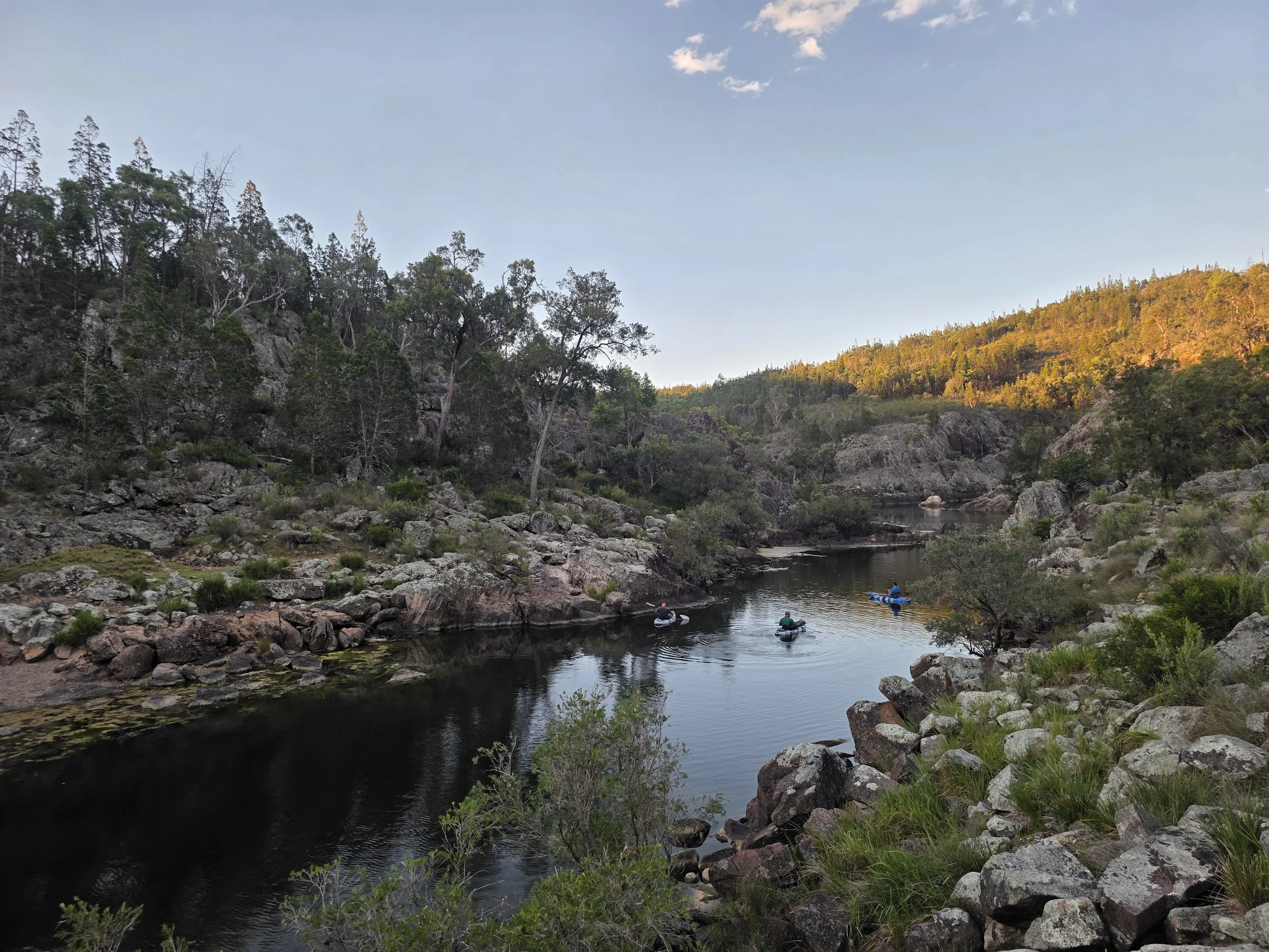 Four people kayaking in a river surrounded by rocky terrain and dense trees in a mountainous landscape during sunset.