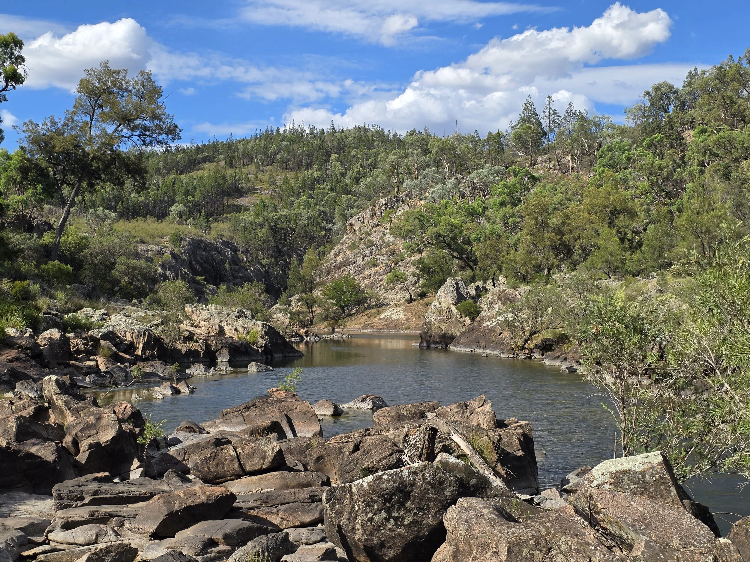 A river flowing through a rocky canyon surrounded by green trees and hills under a partly cloudy blue sky.