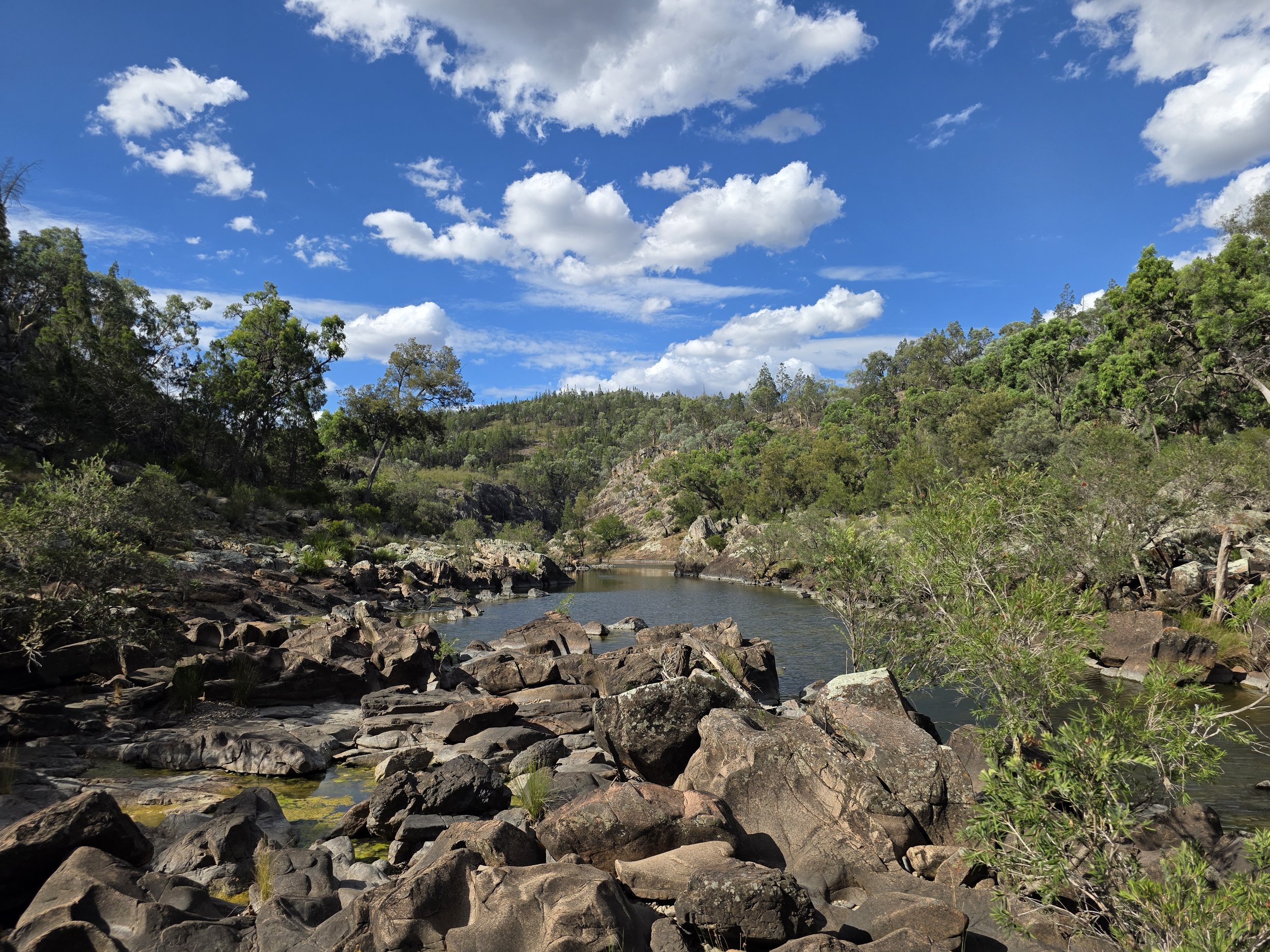 A river flowing through a rocky landscape surrounded by green trees under a blue sky with white clouds.