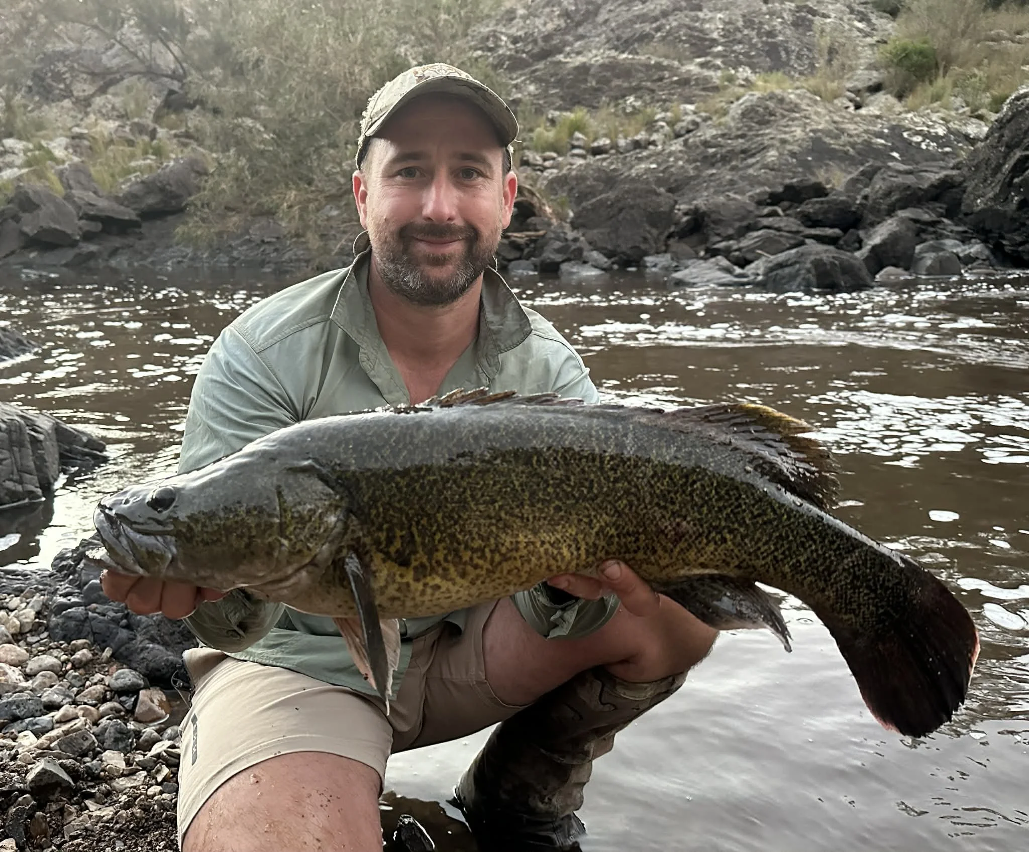 A man with a beard, wearing a cap, green shirt, and beige shorts, kneeling by a river shoreline, holding a large fish and smiling at the camera. The river has rocks and in the background are rocky hills and sparse vegetation.
