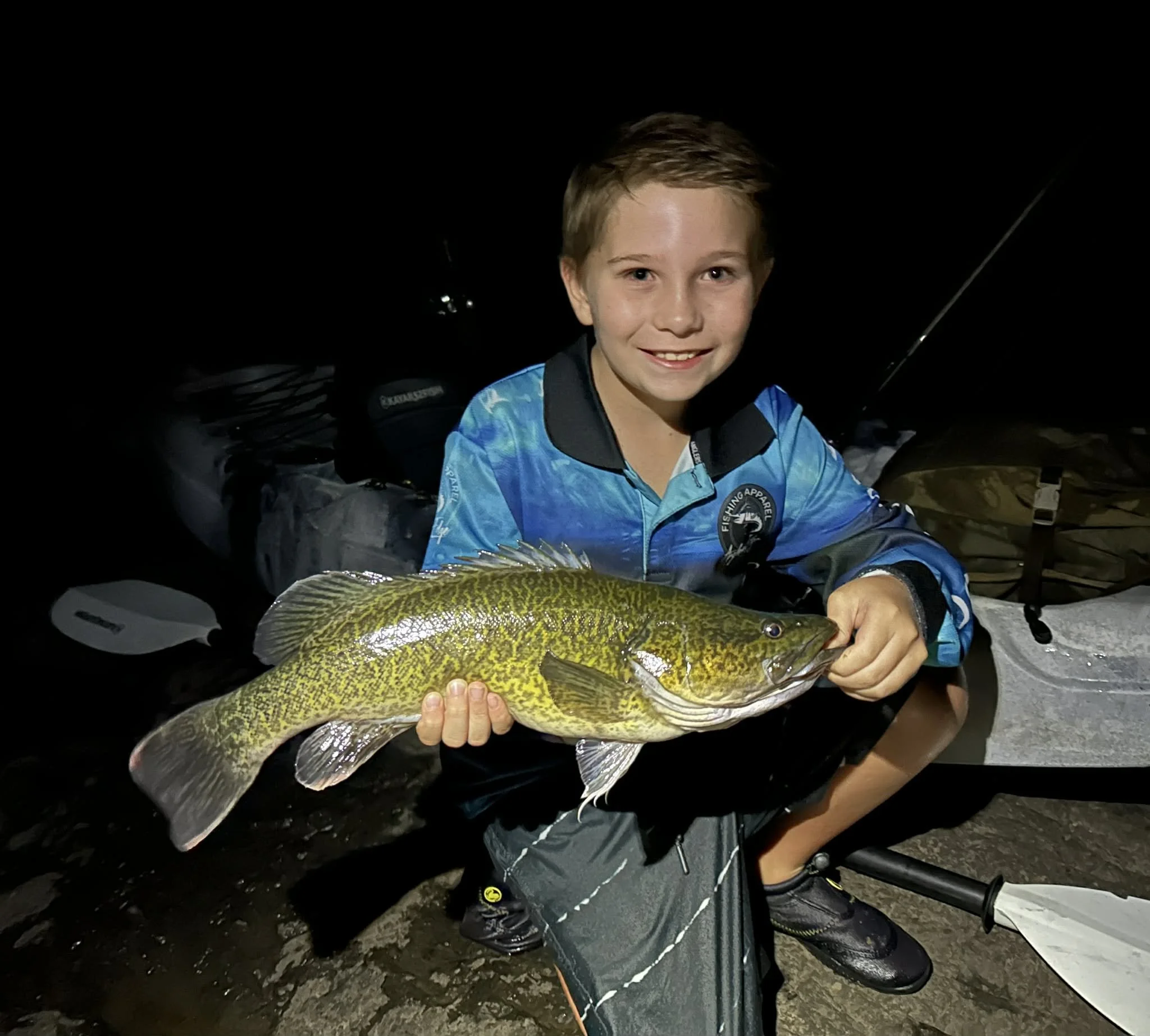 A young boy smiling at night while holding a large fish he caught, with fishing gear and a kayak in the background.