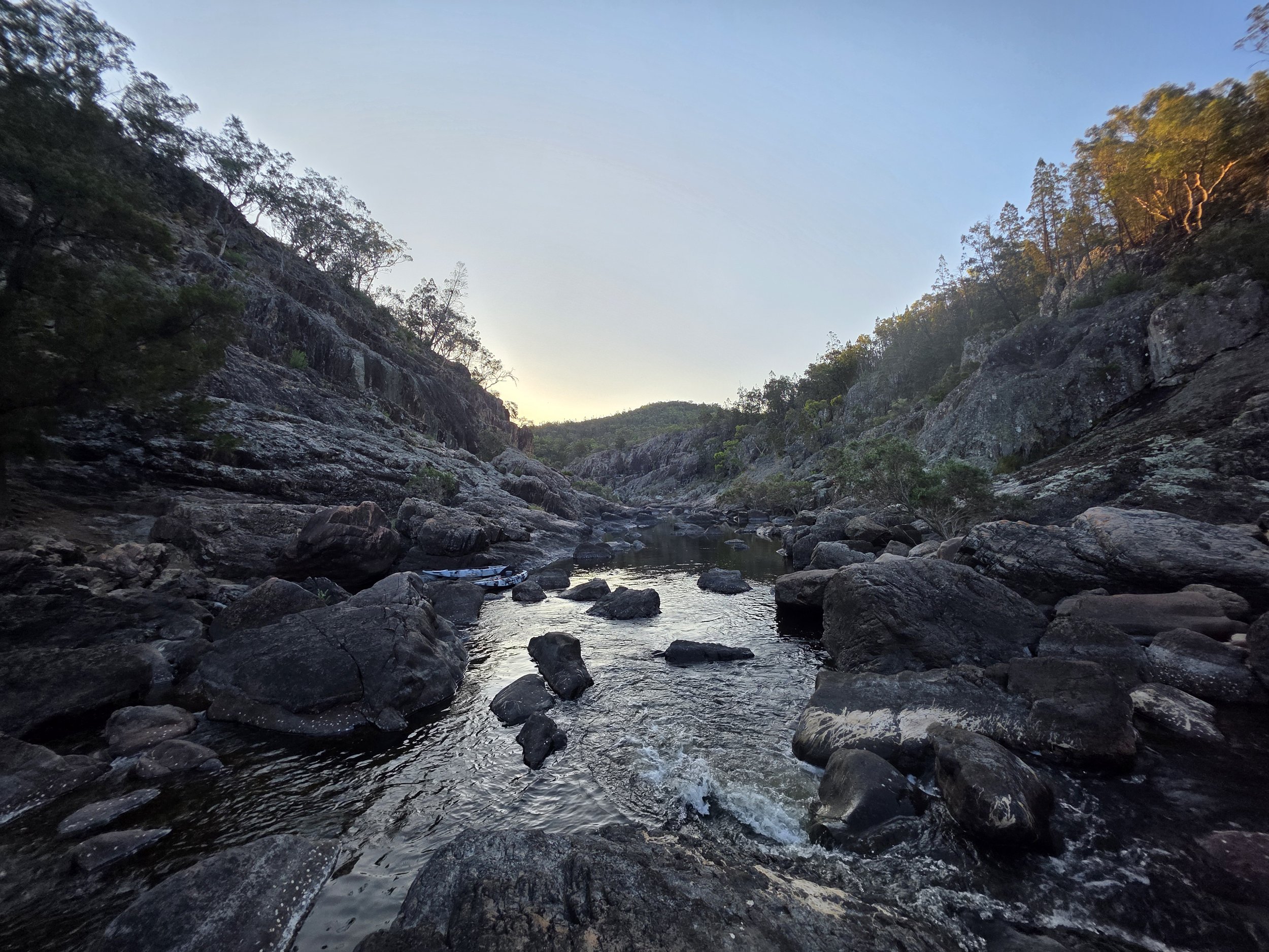 A rocky river surrounded by trees and hills, with the sky visible at sunset or sunrise.