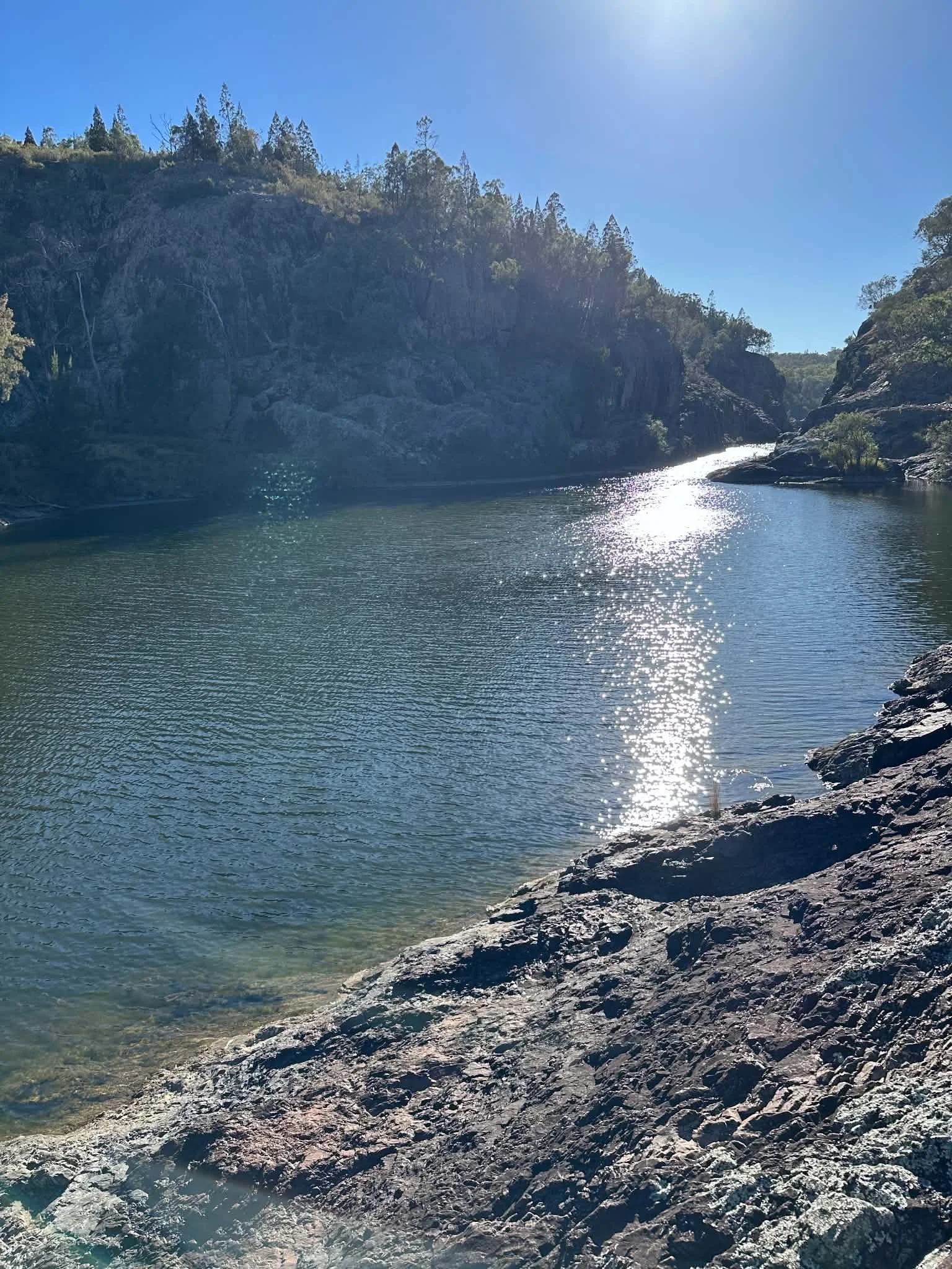 A scenic view of a river flowing through rocky terrain with hills covered in trees under a clear blue sky, sunlight reflecting off the water.
