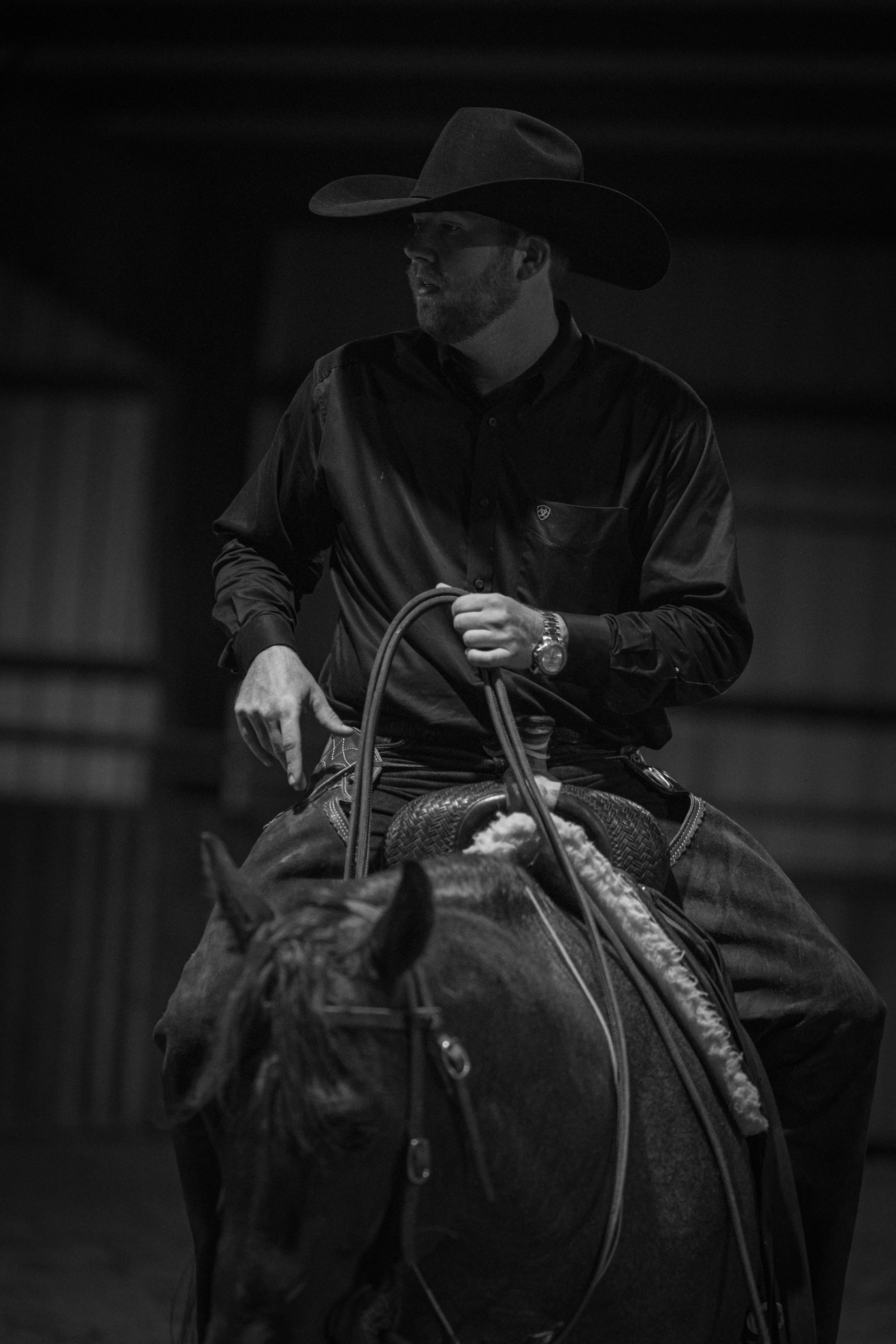 A man wearing a cowboy hat and a long-sleeve shirt riding a horse in a dark setting.