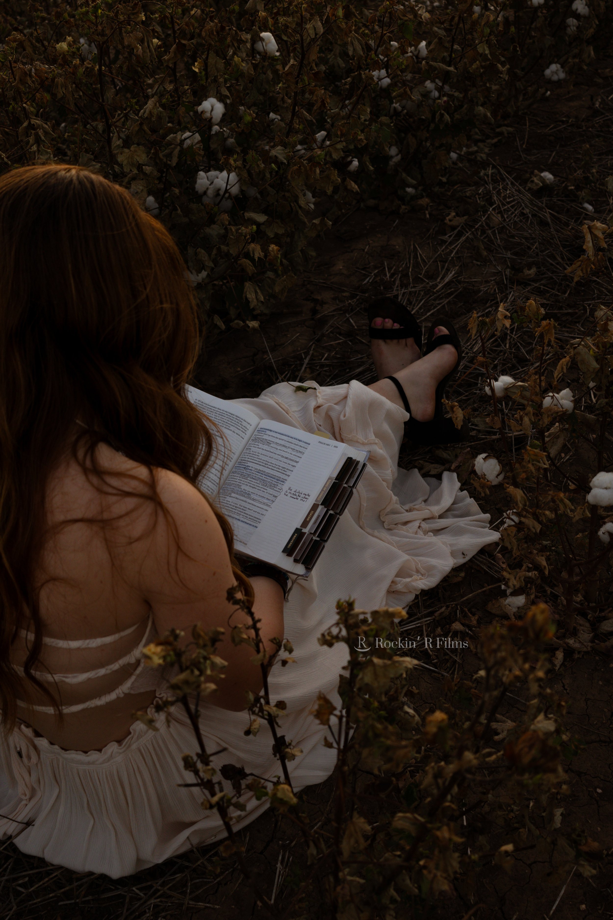 A woman with long red hair seated on dry soil among cotton plants, reading a book with black markers on her lap, wearing a light-colored dress and black sandals.