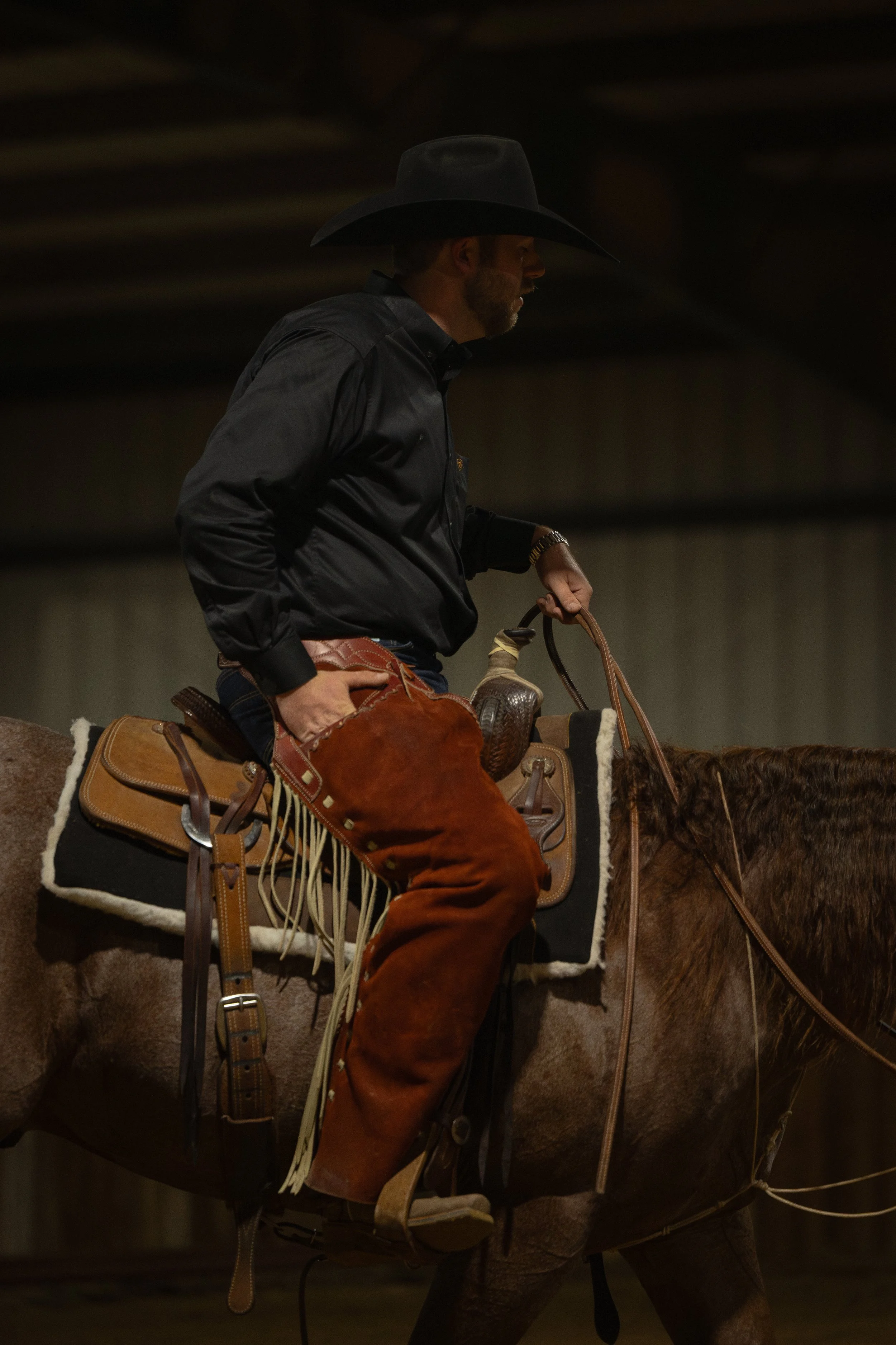 A cowboy riding a horse indoors, wearing a black cowboy hat, black shirt, and brown chaps with fringe, holding the reins with one hand.