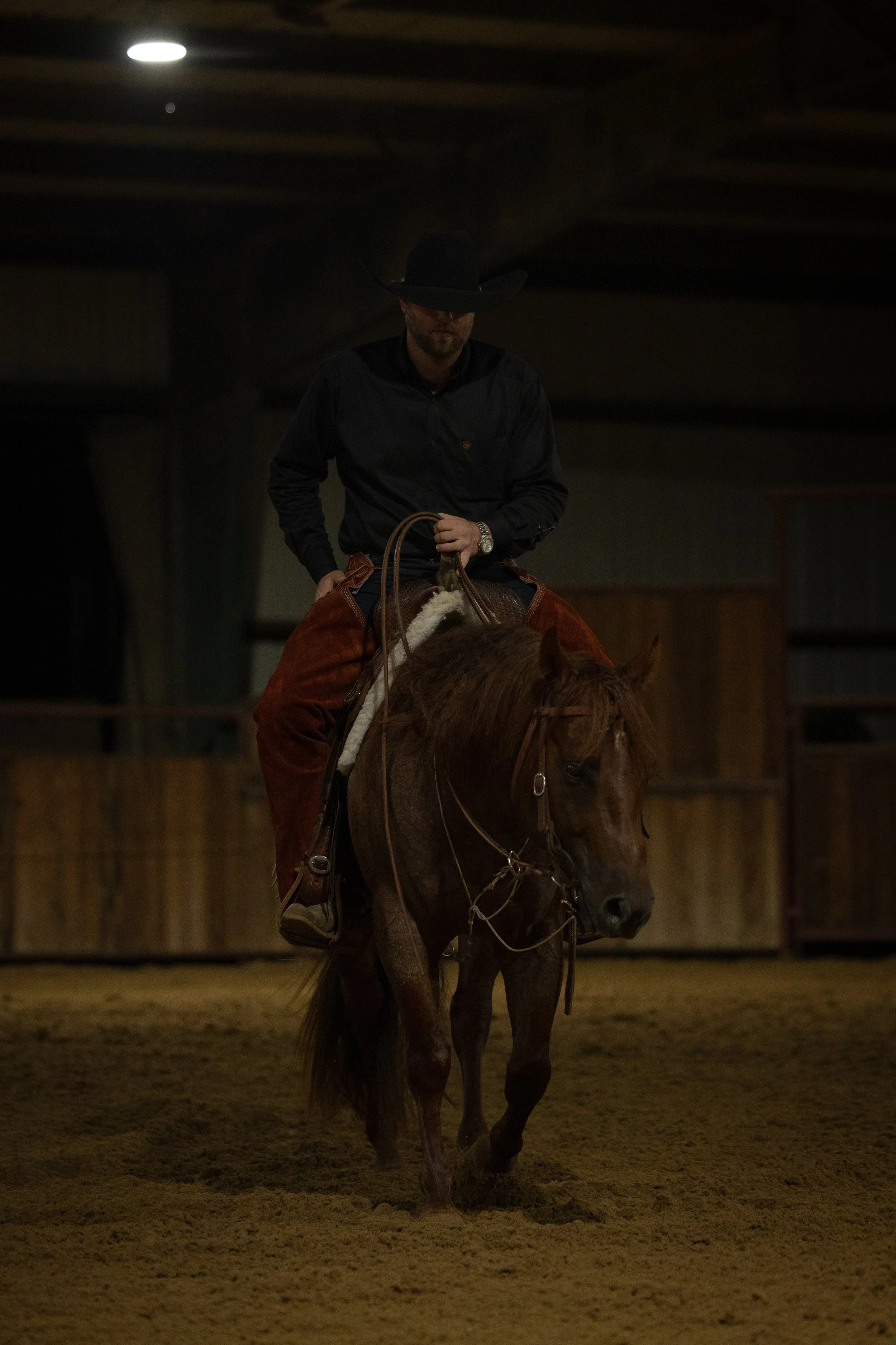 A man riding a horse indoors, wearing a black cowboy hat, black shirt, and brown chaps.