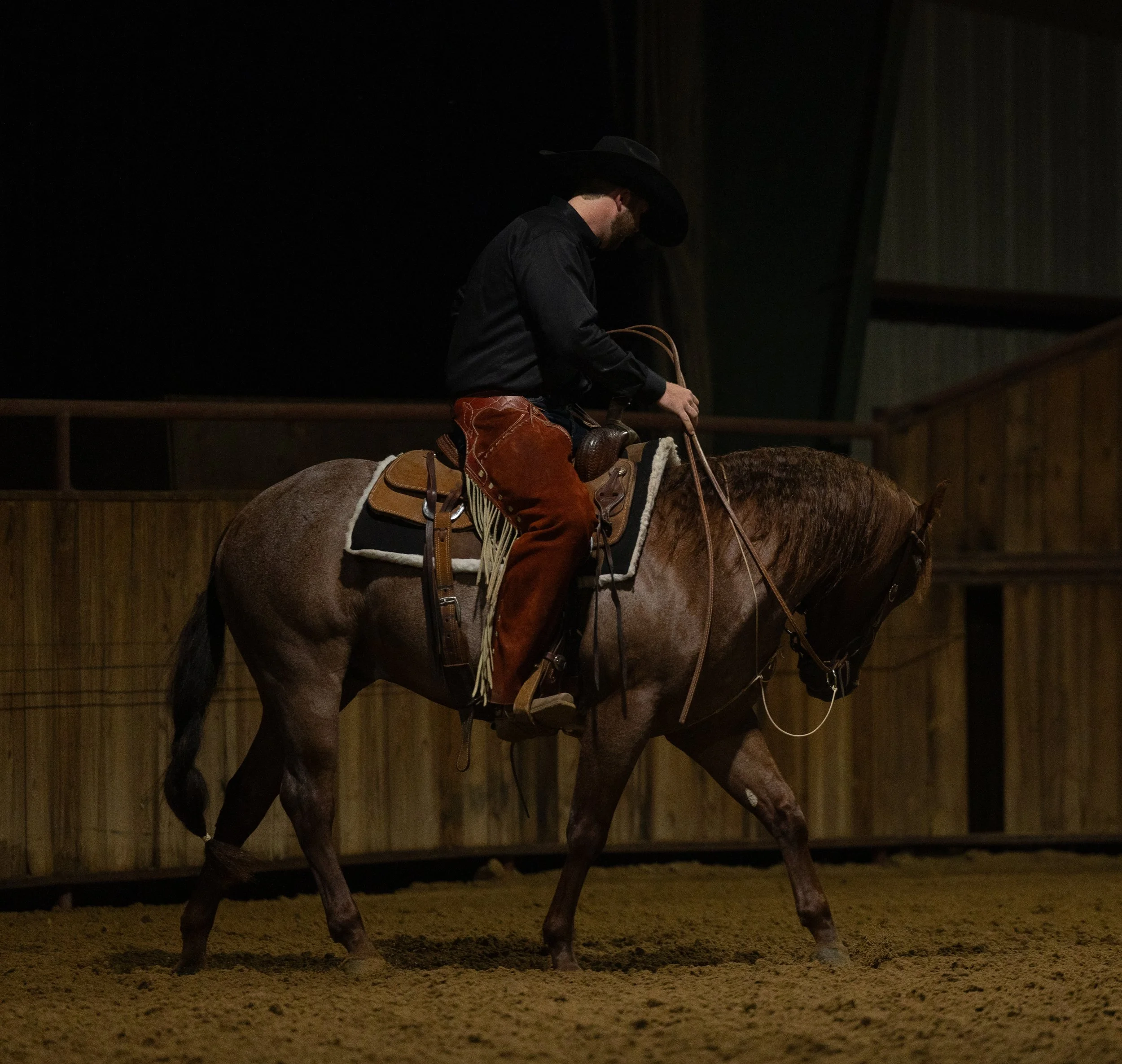 A man dressed in Western attire, including a black cowboy hat, black shirt, and brown chaps, riding a brown horse in an indoor arena with wooden fencing.