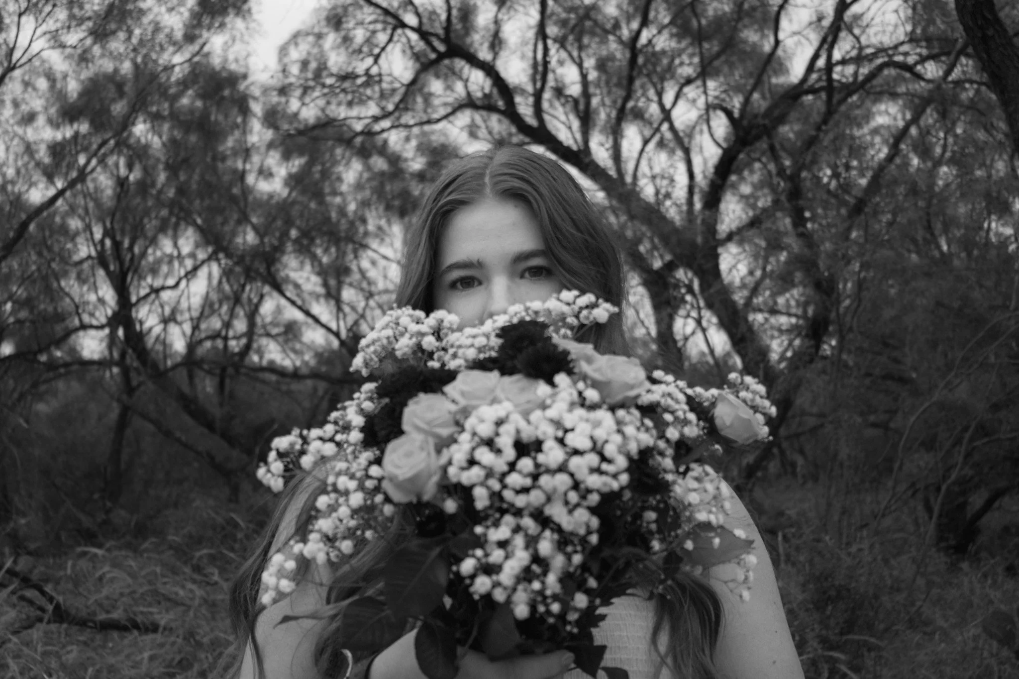 A young woman with long hair holding a bouquet of flowers in front of her face, outdoors with trees in the background, in black and white.