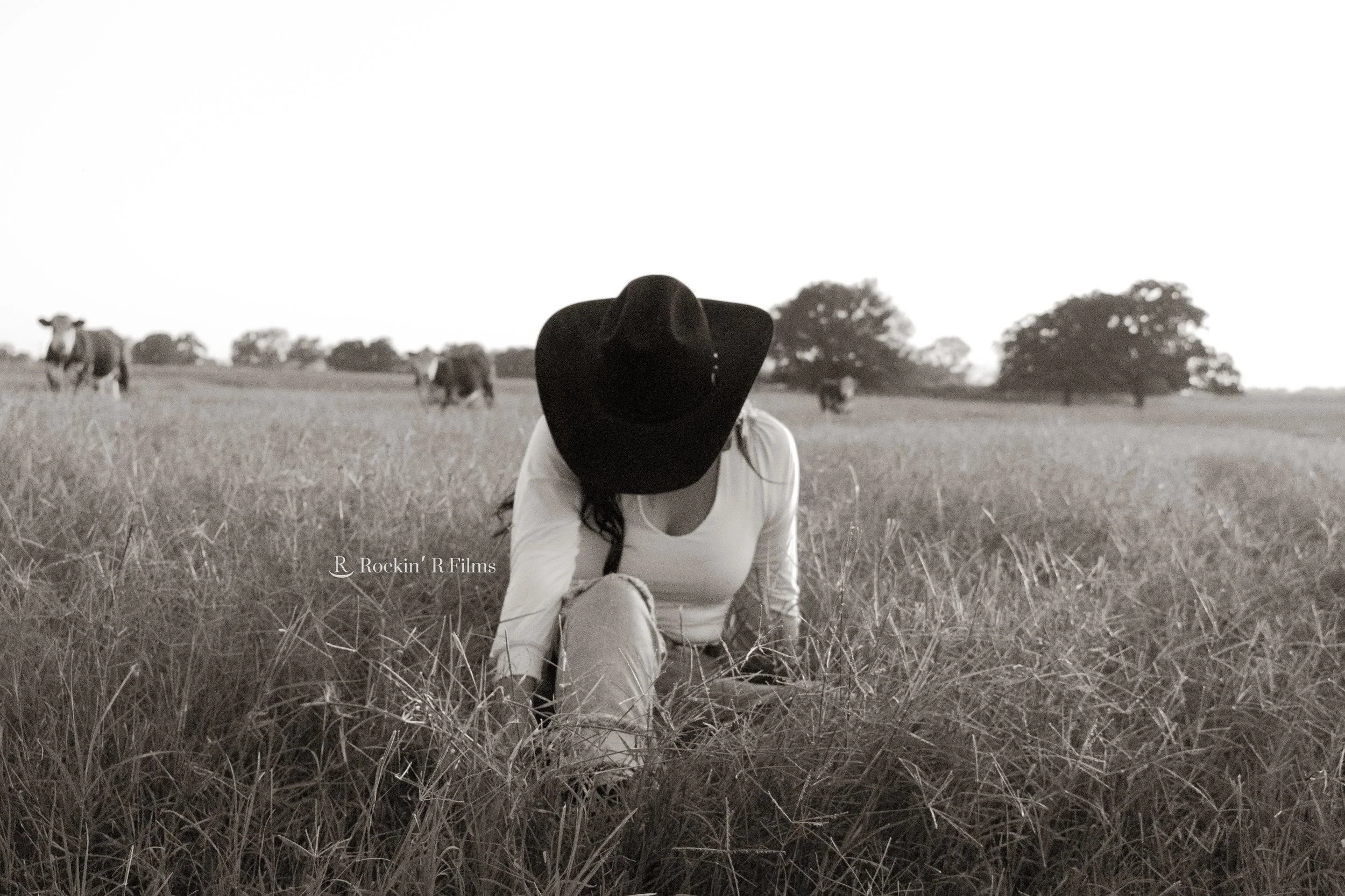 A woman wearing a large black hat and light-colored clothing crouches in a grassy field with cows in the background.