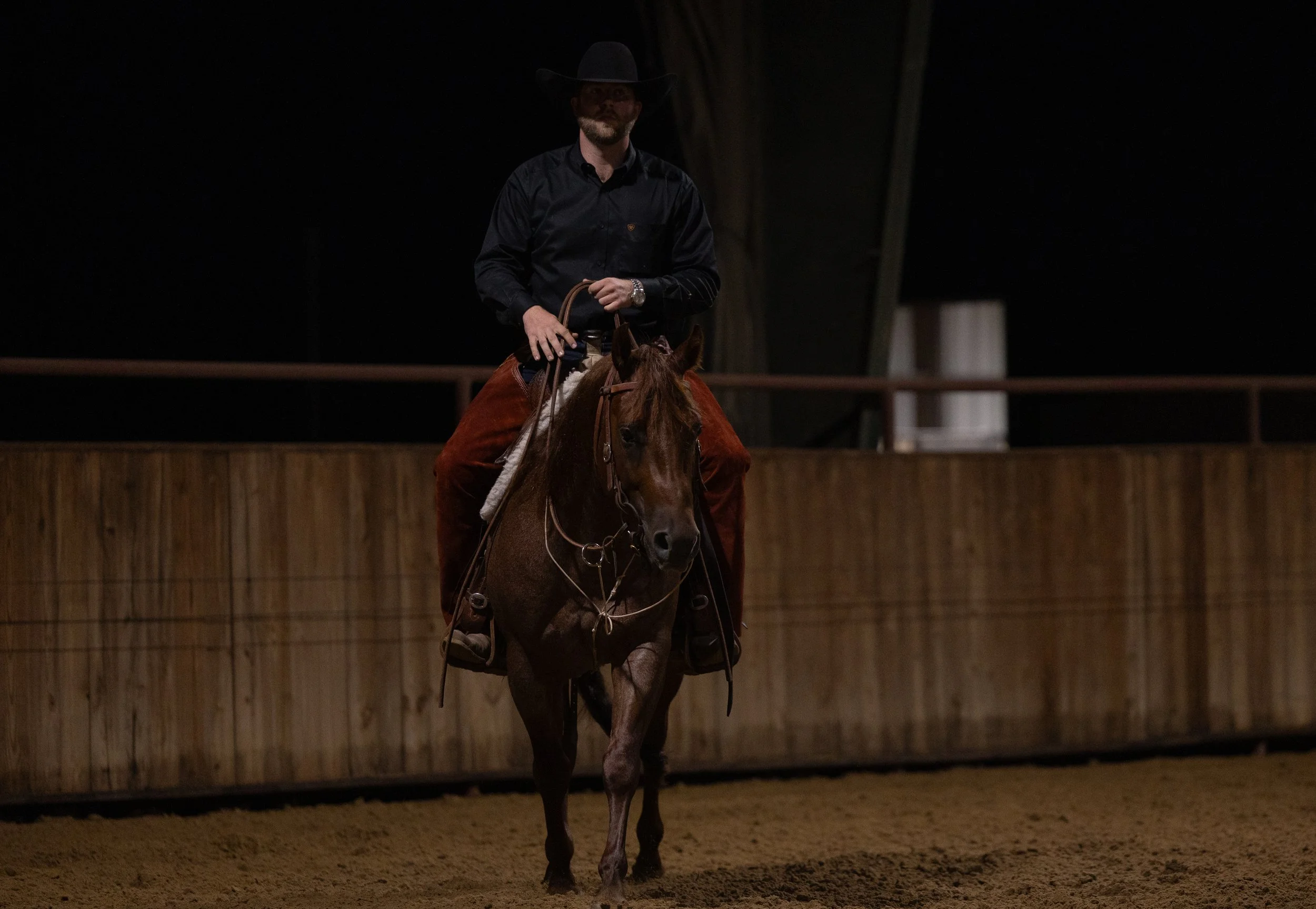 A man wearing a black cowboy hat, black shirt, and brown pants riding a brown horse inside a wooden-fenced indoor arena at night.