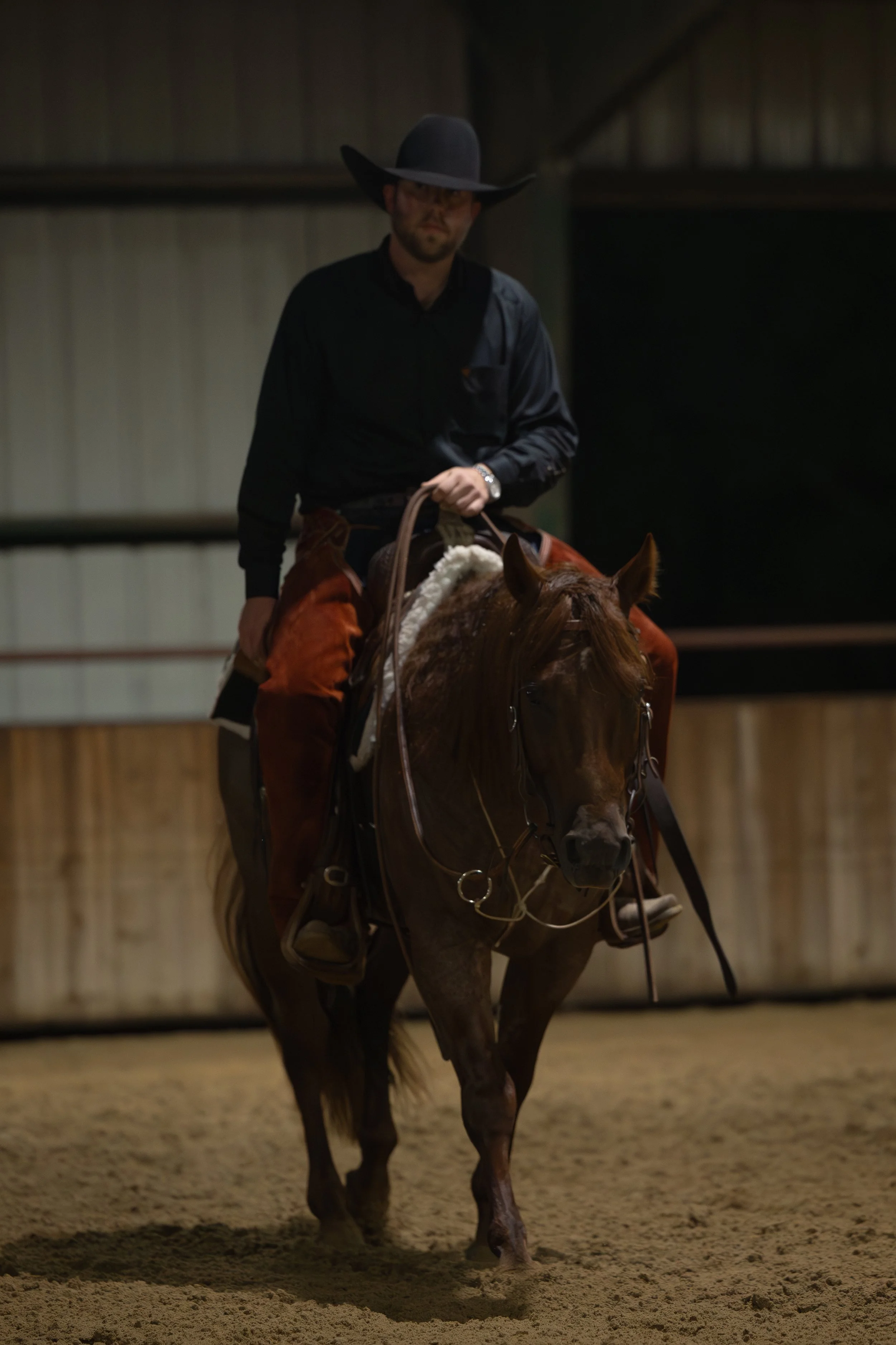A man wearing a black shirt, brown chaps, and a black cowboy hat riding a brown horse inside an indoor arena.