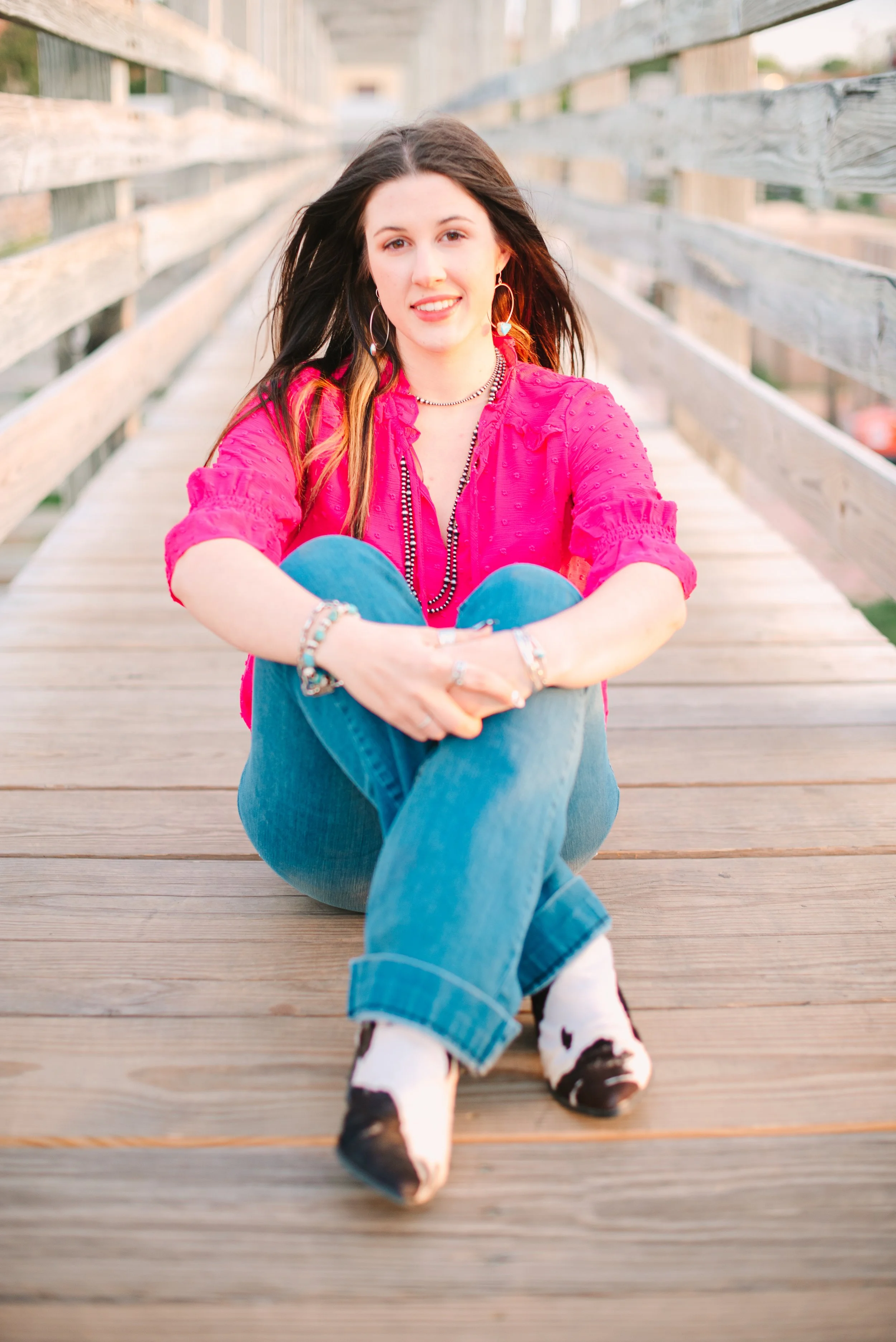 A young woman with dark brown hair, wearing a bright pink blouse and blue jeans, sitting on a wooden bridge with her arms wrapped around her knees and smiling at the camera.