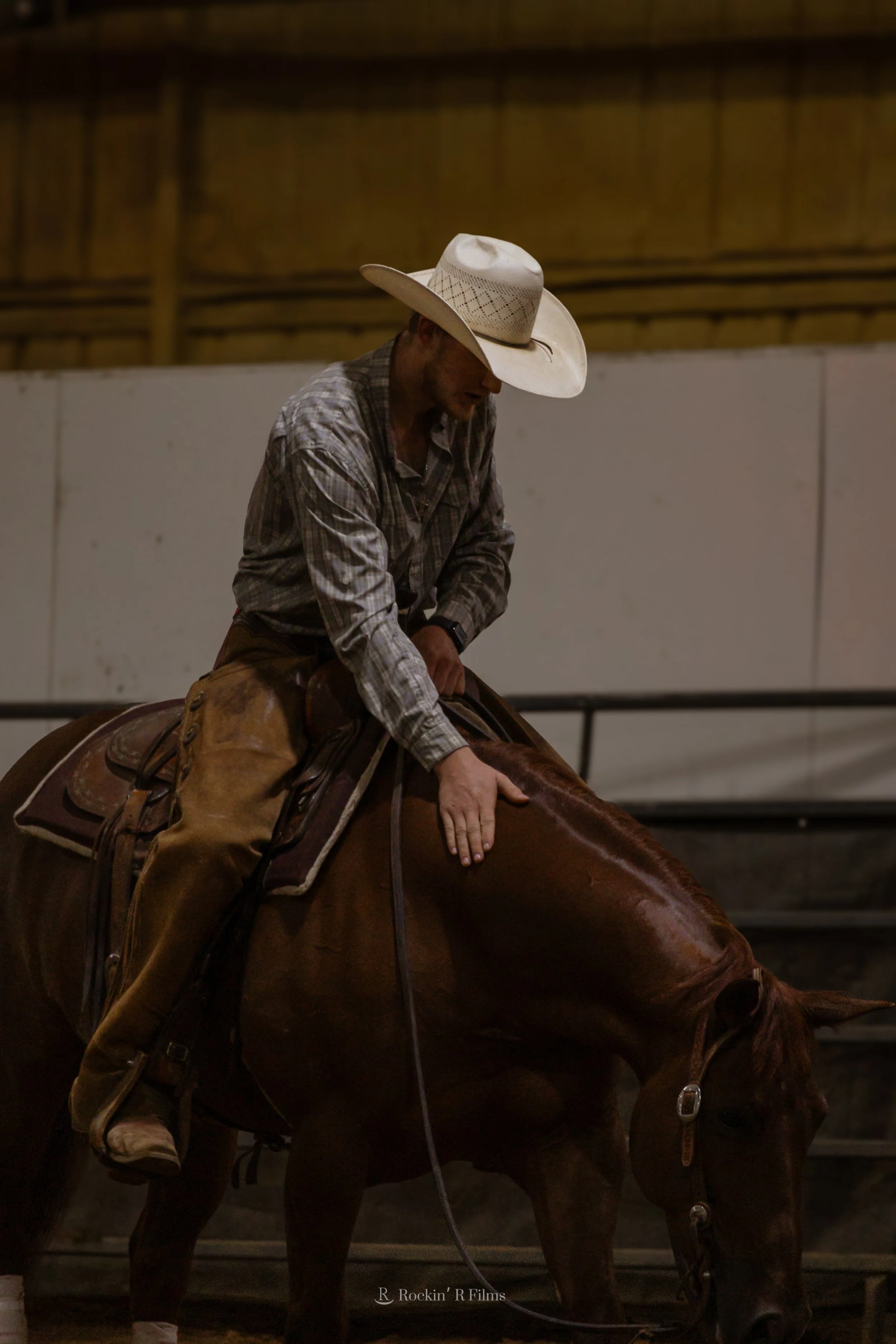 A man wearing a cowboy hat, plaid shirt, and leather chaps riding a brown horse inside an indoor arena.