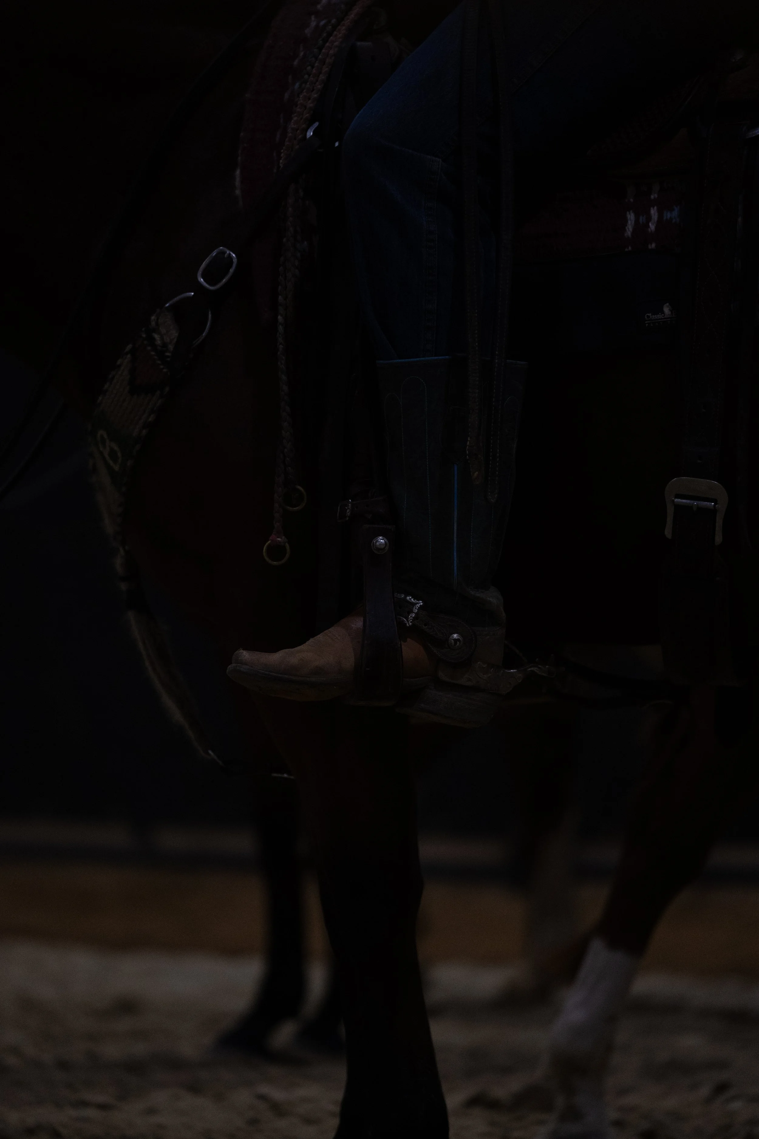 A horse with a saddle and riding gear, standing on dirt ground, with legs and hooves visible.