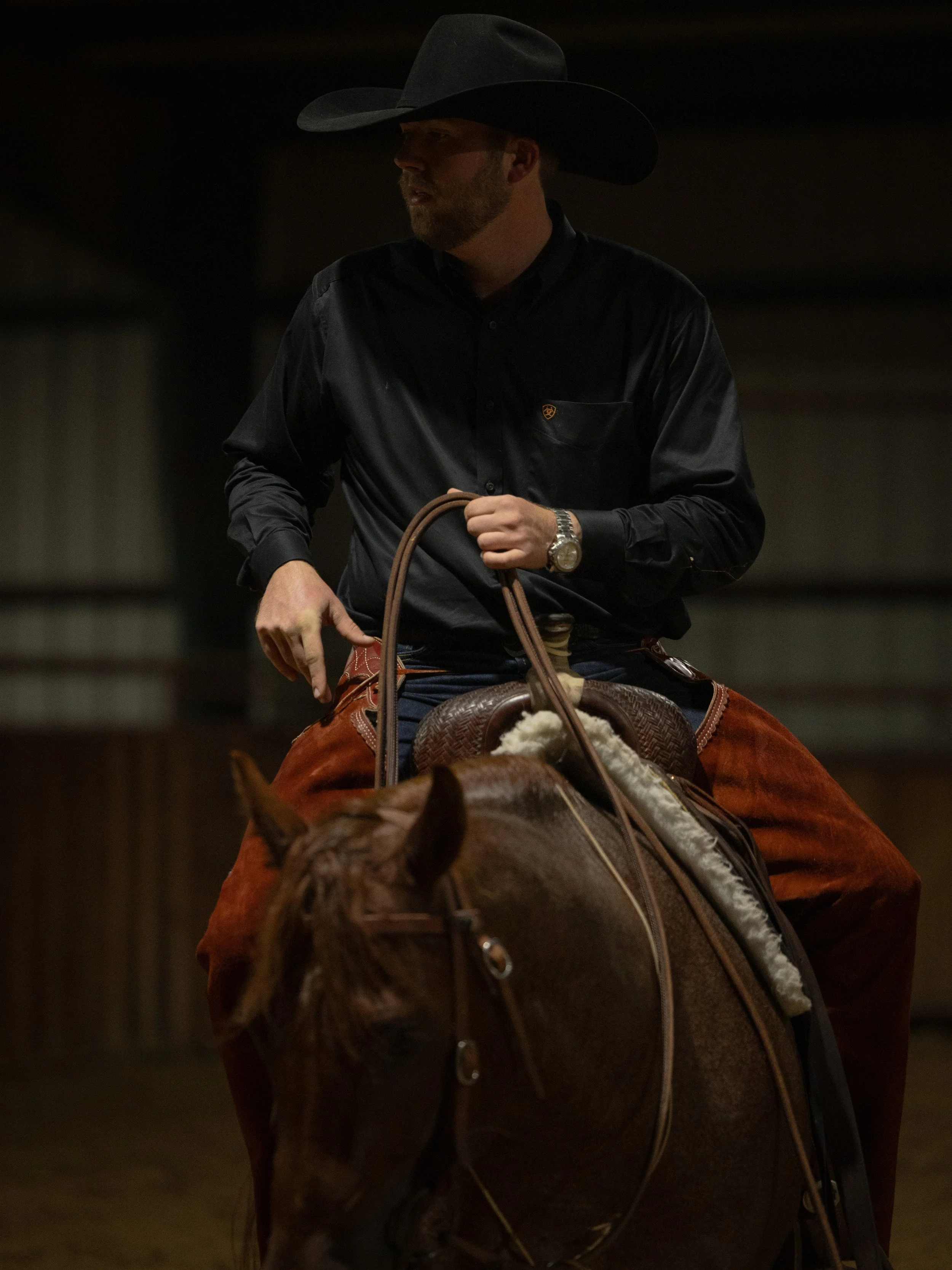 A man riding a horse in an indoor riding arena, wearing a black cowboy hat, a black button-up shirt, and brown chaps.