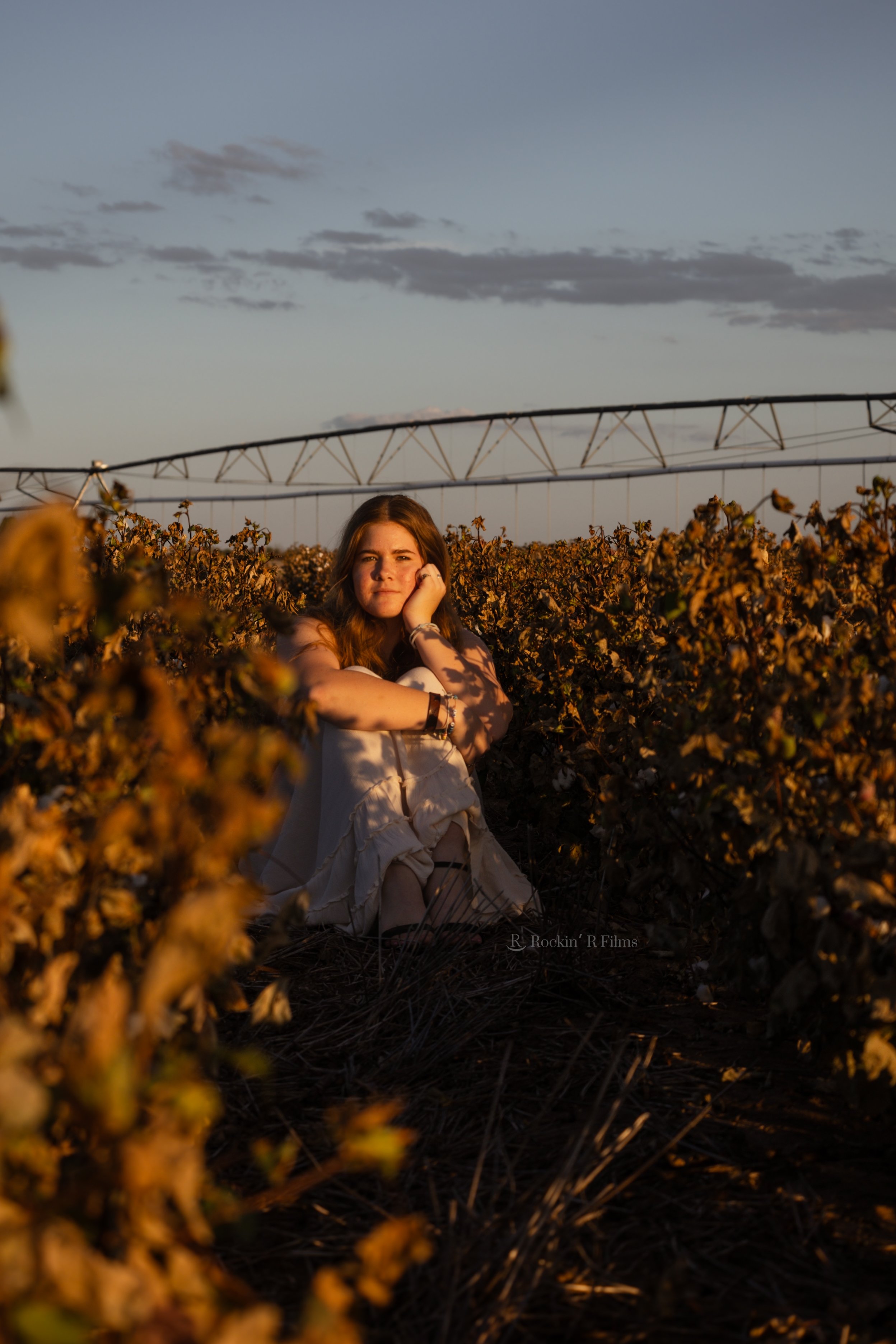 A young woman with long brown hair sitting in a vineyard at sunset, surrounded by grapevines, with a distant sky and a wind turbine in the background.