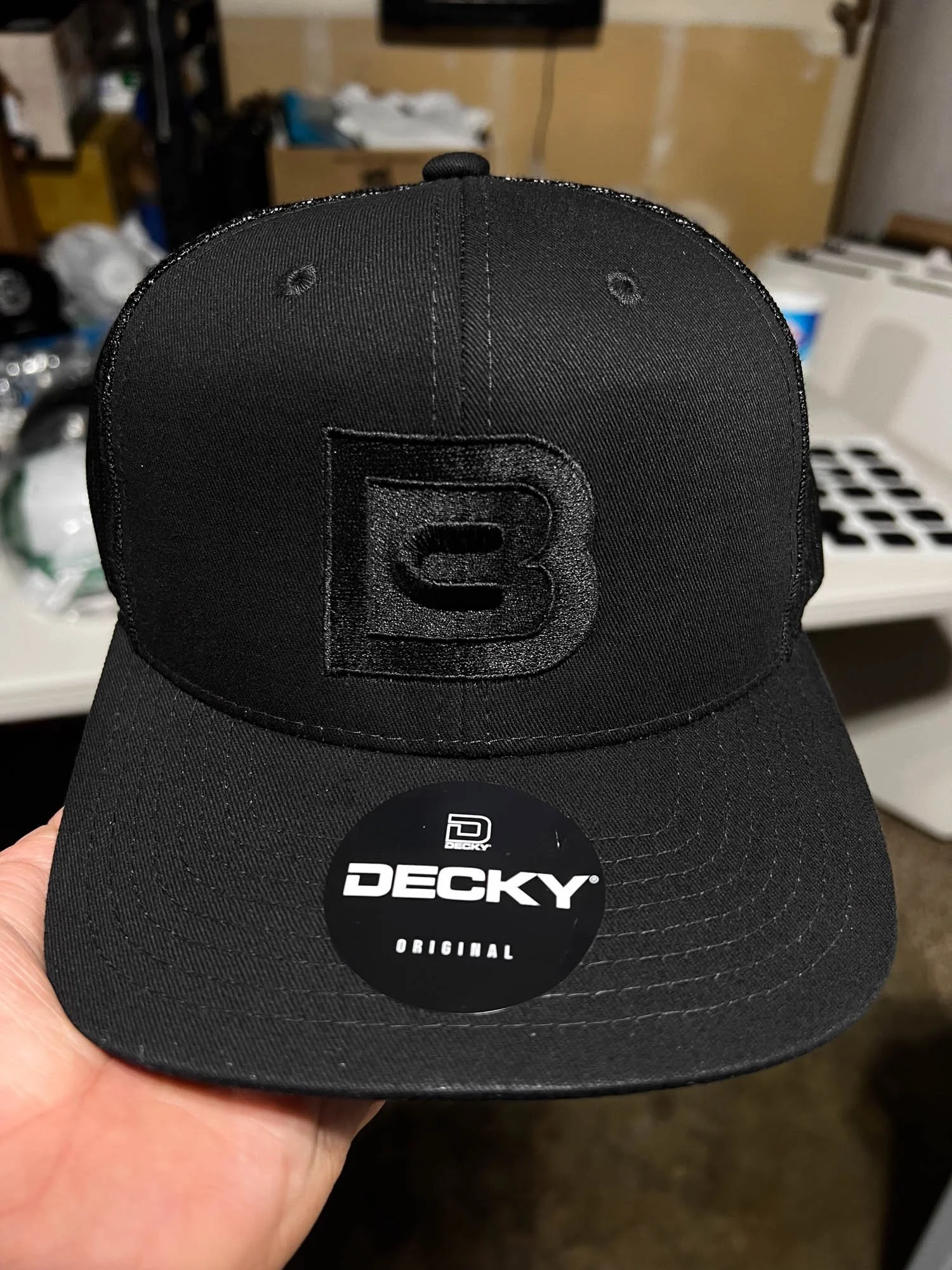 Black DECKY baseball cap with an embroidered logo on the front, held in a person's hand, with a background of miscellaneous items and a washer and dryer.