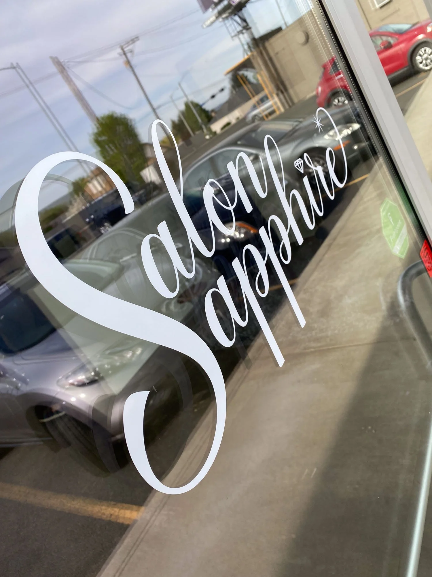 Close-up of a glass door with white cursive text that reads "Salon Saphi" and a small diamond icon, reflecting parking lot and cars outside.