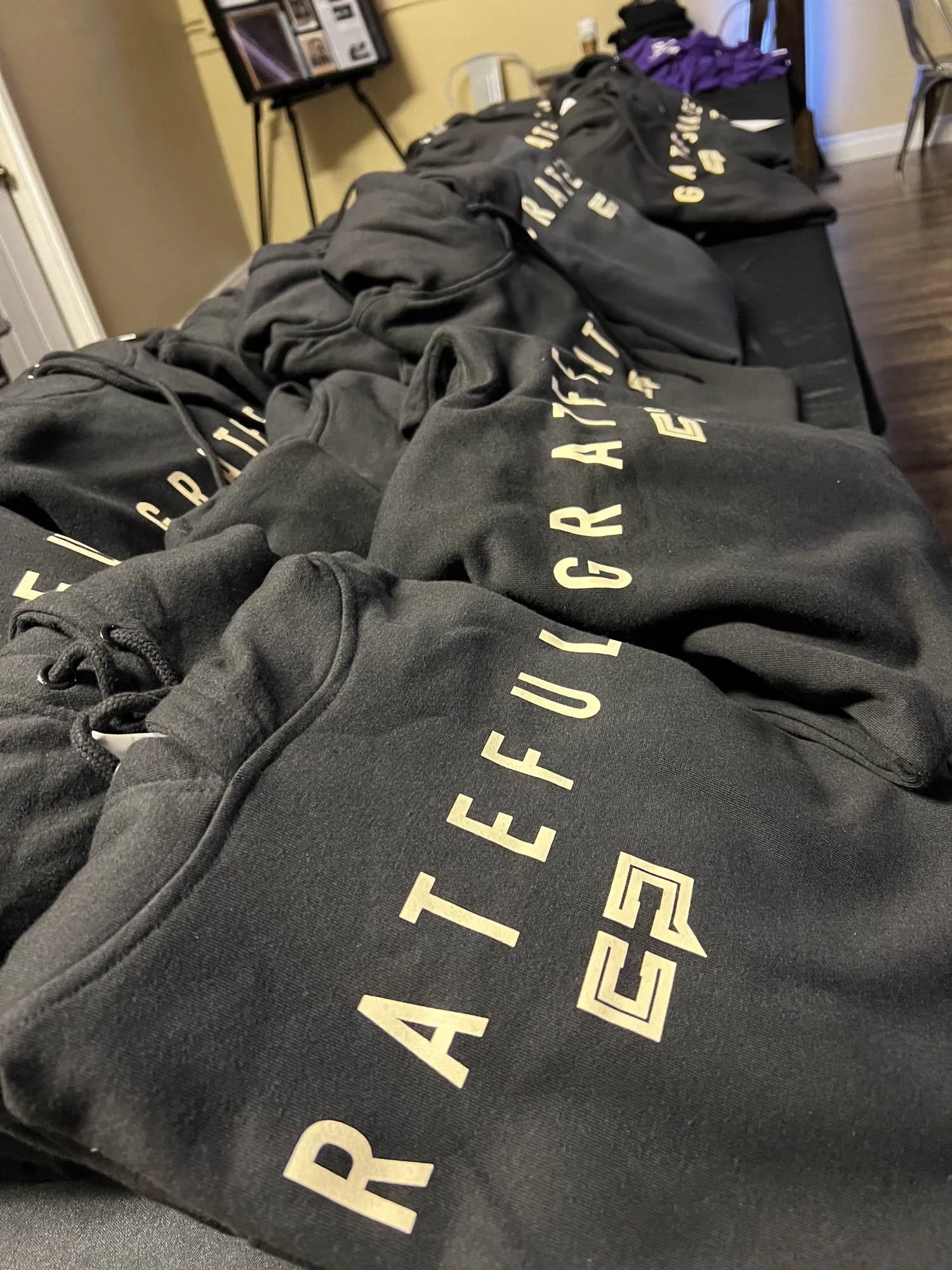 Black hoodies with 'RAIDER ARTIST' printed in gold lettering, laid out on a long table indoors.