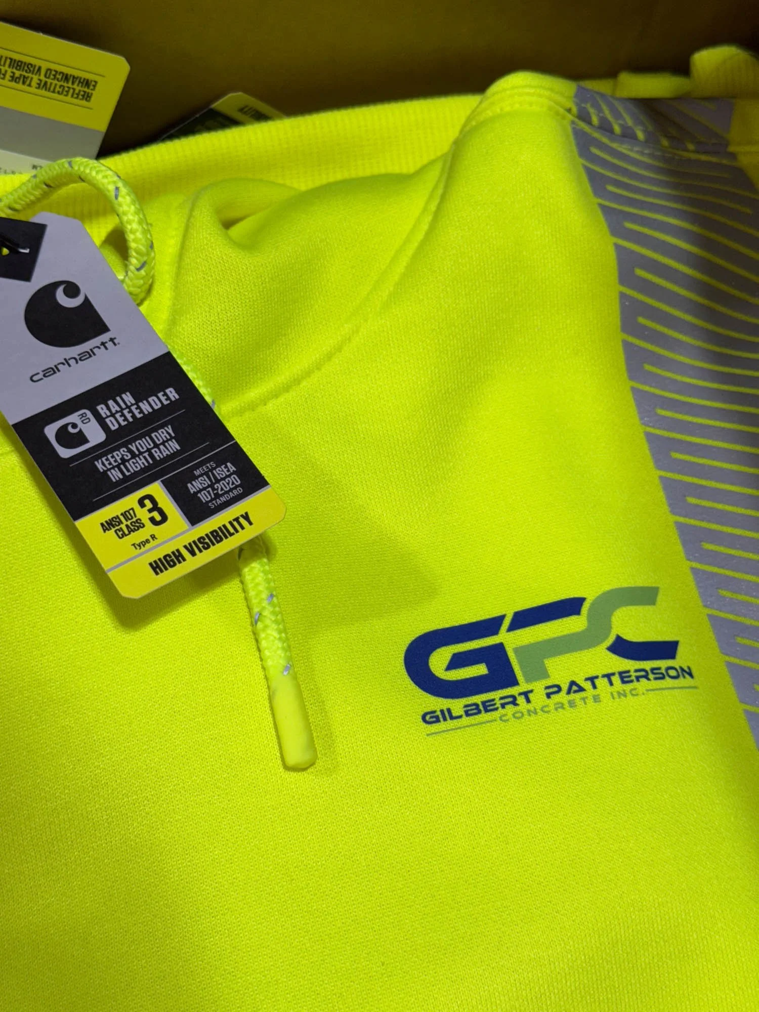 Close-up of a yellow high-visibility rain jacket with a zipper, featuring the logo and name of Gilbert Patterson Concrete Inc. and a Carhartt tag indicating high visibility and rain protection.