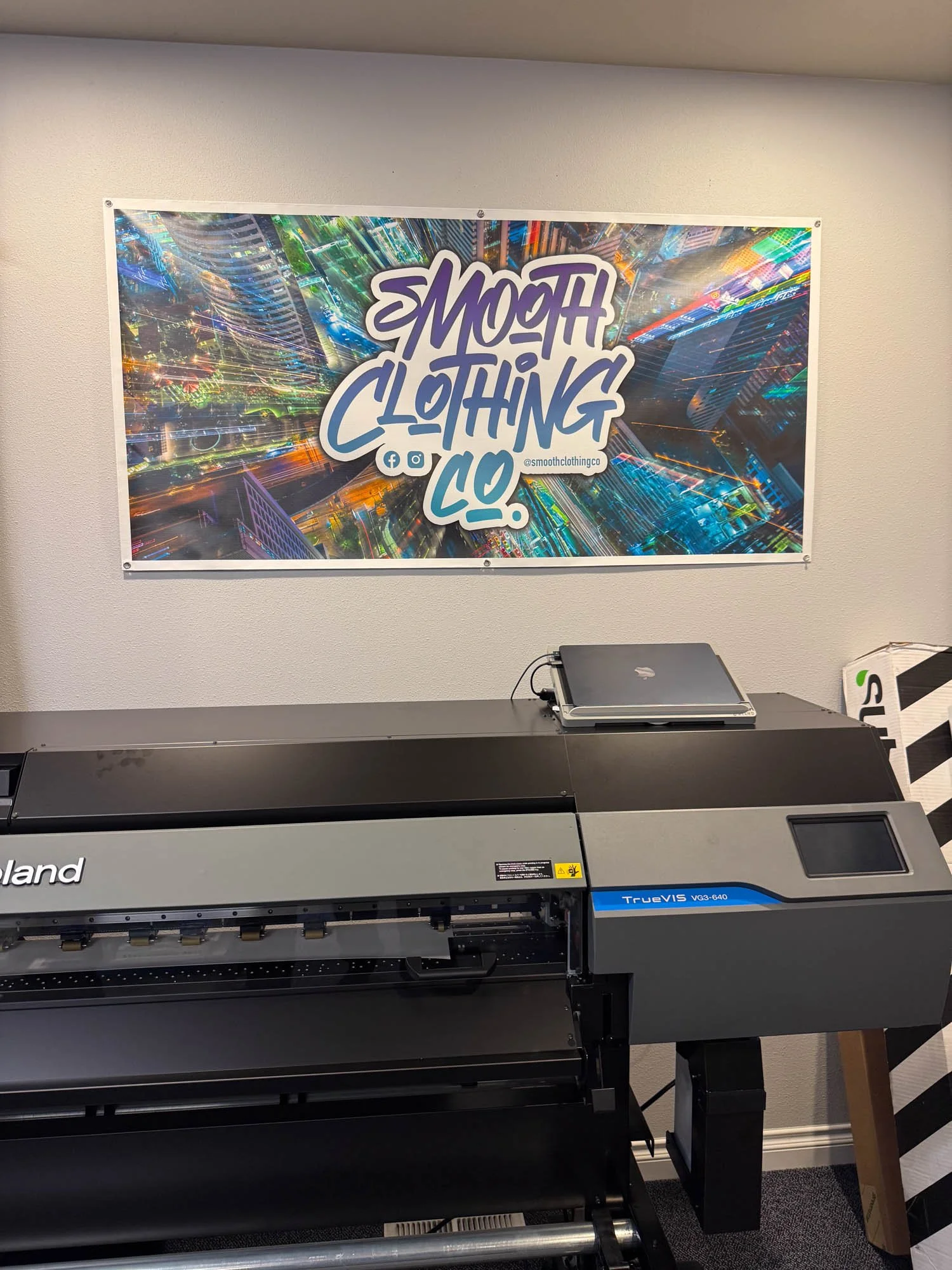 A large flatbed printing machine in front of a wall with a colorful urban cityscape banner promoting 'Smooth Clothing Co.' with social media icons and handle '@smoothclothingco'.