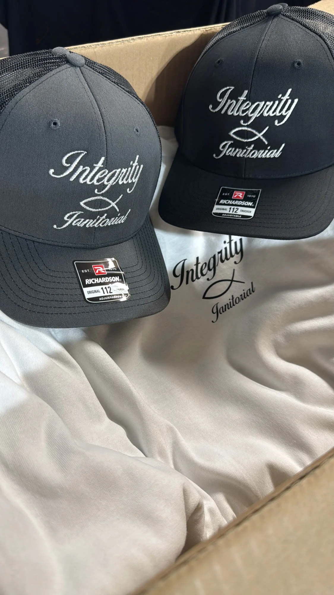 Two black baseball caps with embroidered white text reading 'Integrity Tanitual' are placed inside a cardboard box with cream-colored fabric lining. There is also a cloth with the same text.
