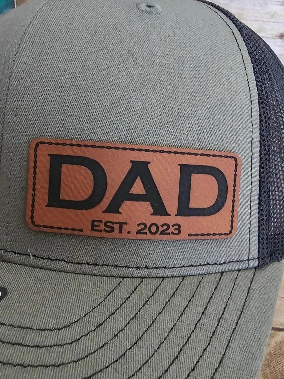 A gray cap with a leather patch that reads "DAD EST. 2023" in black letters.
