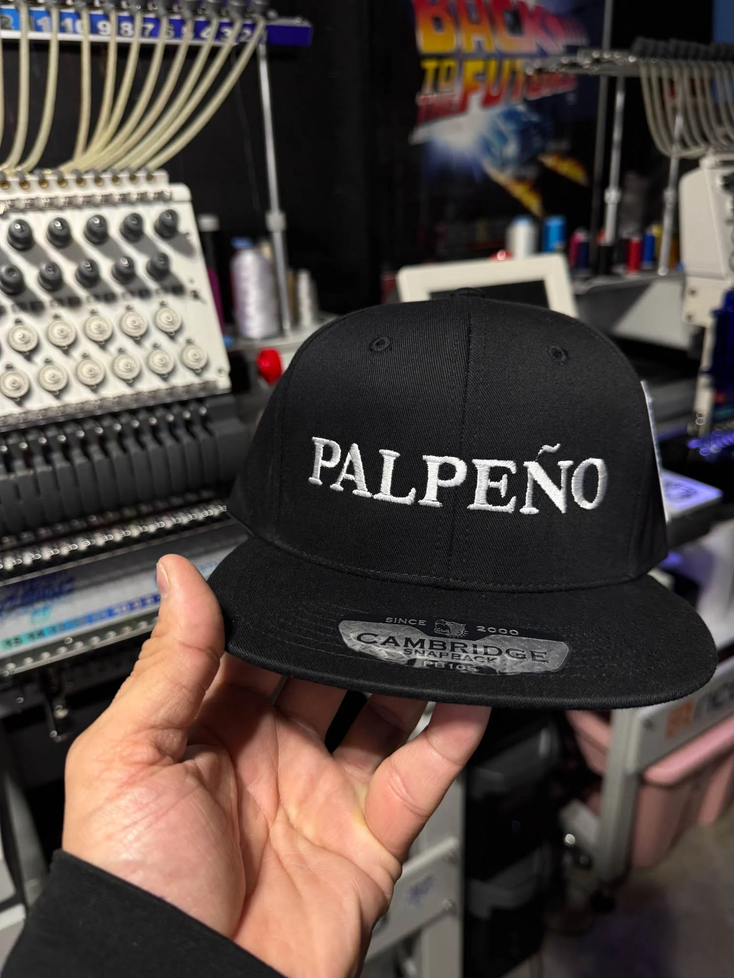 A person holding a black baseball cap with the embroidered word 'PALPEÑO' on the front. The background shows sewing or embroidery machines and supplies in a workshop setting.