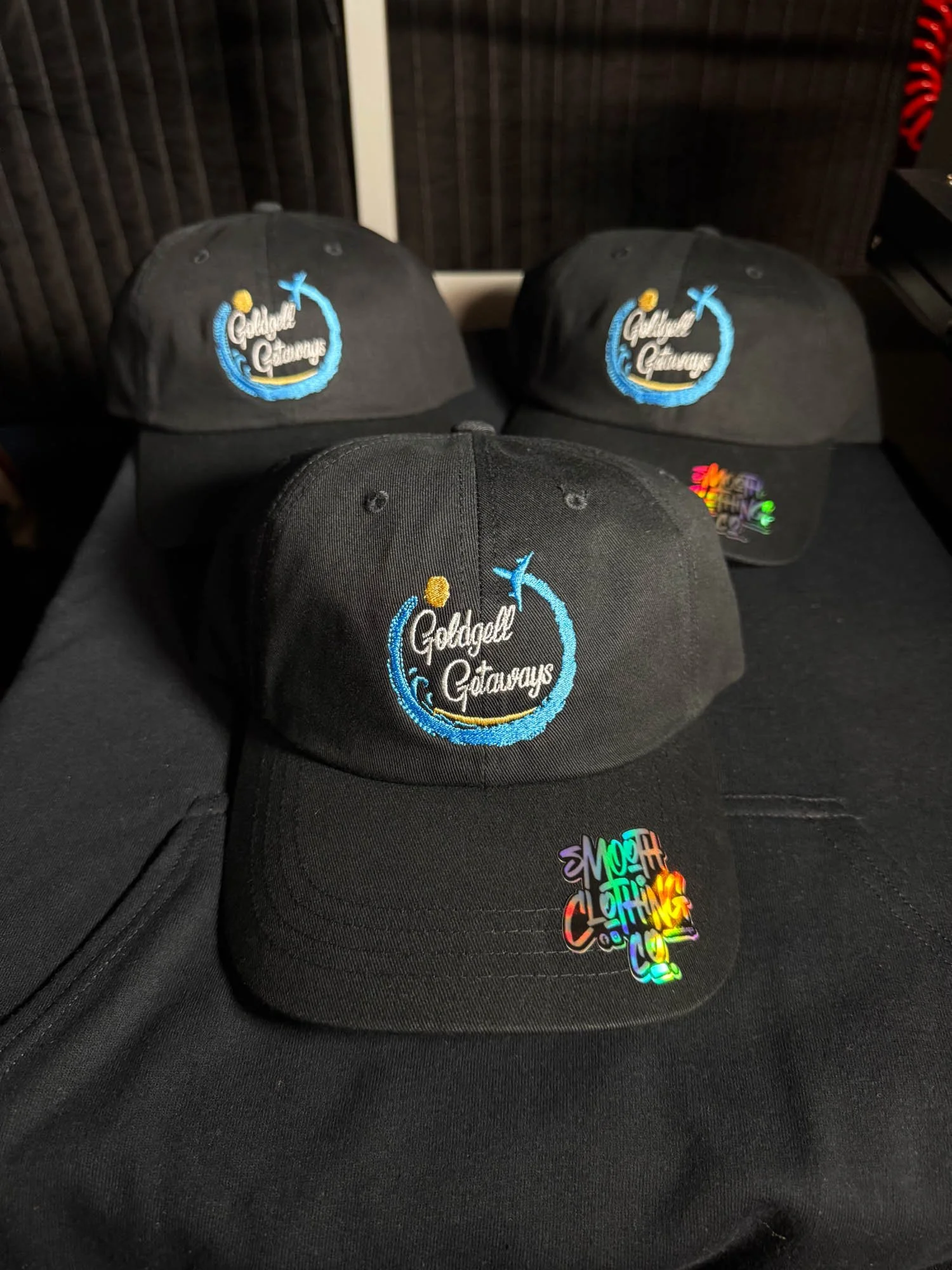 Four black baseball caps with embroidered logos that say 'Gilded Getaways' with a blue wave and yellow dot, placed on a black table.