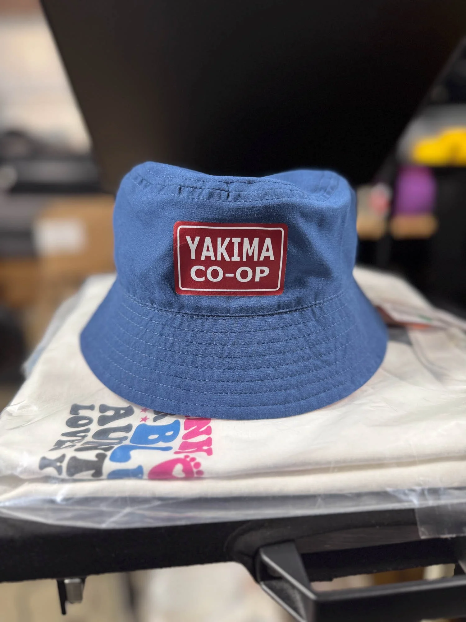 A blue bucket hat with a red and white label reading 'YAKIMA CO-OP' sitting on a plastic bag on a black table.