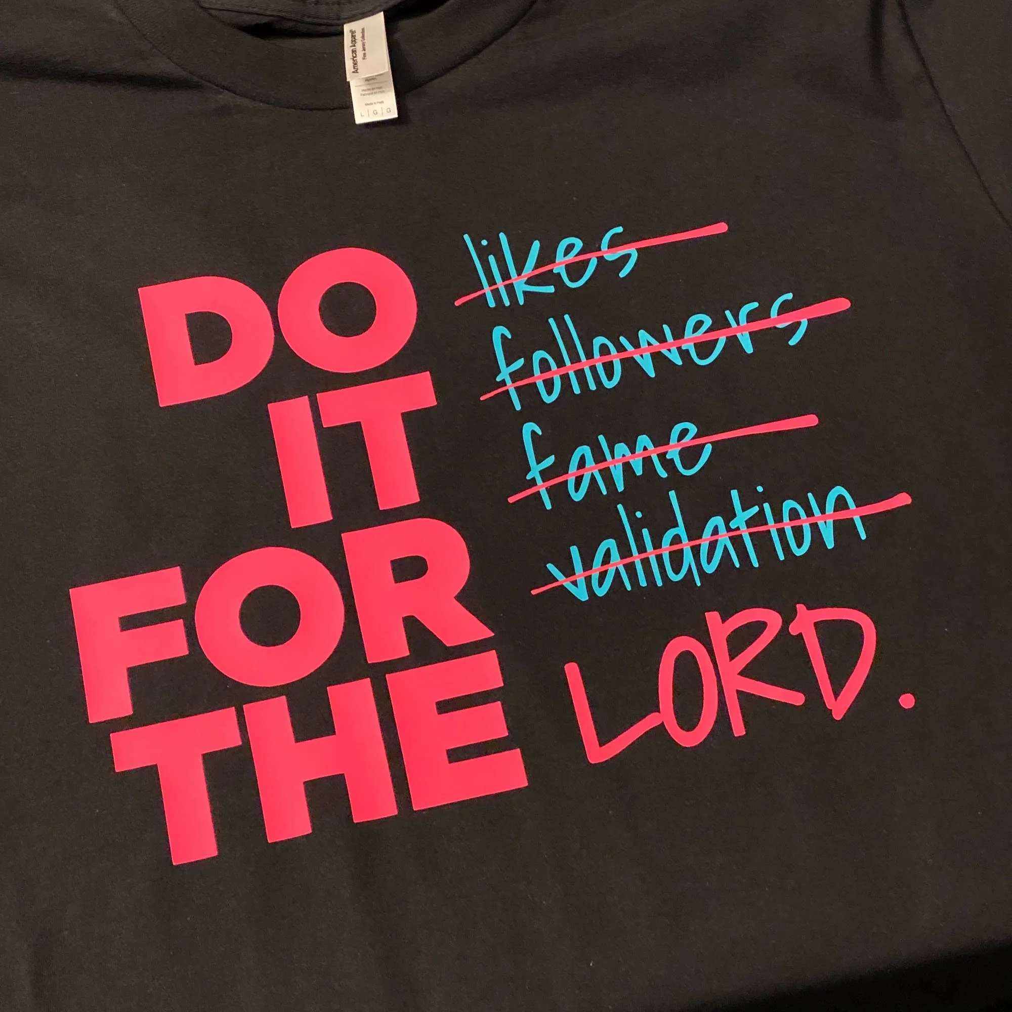 A black T-shirt with pink and blue text. The large pink text reads 'DO IT FOR THE' and the blue words 'likes,' 'followers,' 'fame,' and 'validation' are crossed out in pink, with the phrase 'LORD' written in pink at the bottom.