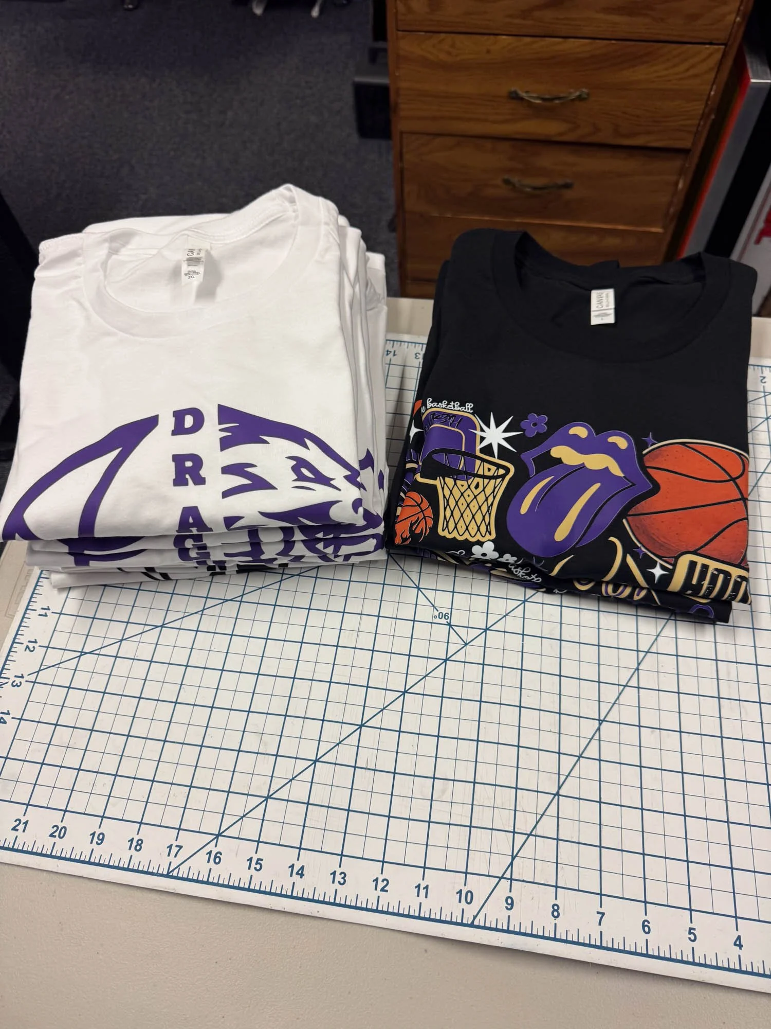 Two folded t-shirts on a cutting mat, one white with purple Baltimore Ravens logo and one black with colorful sports-themed graphics.