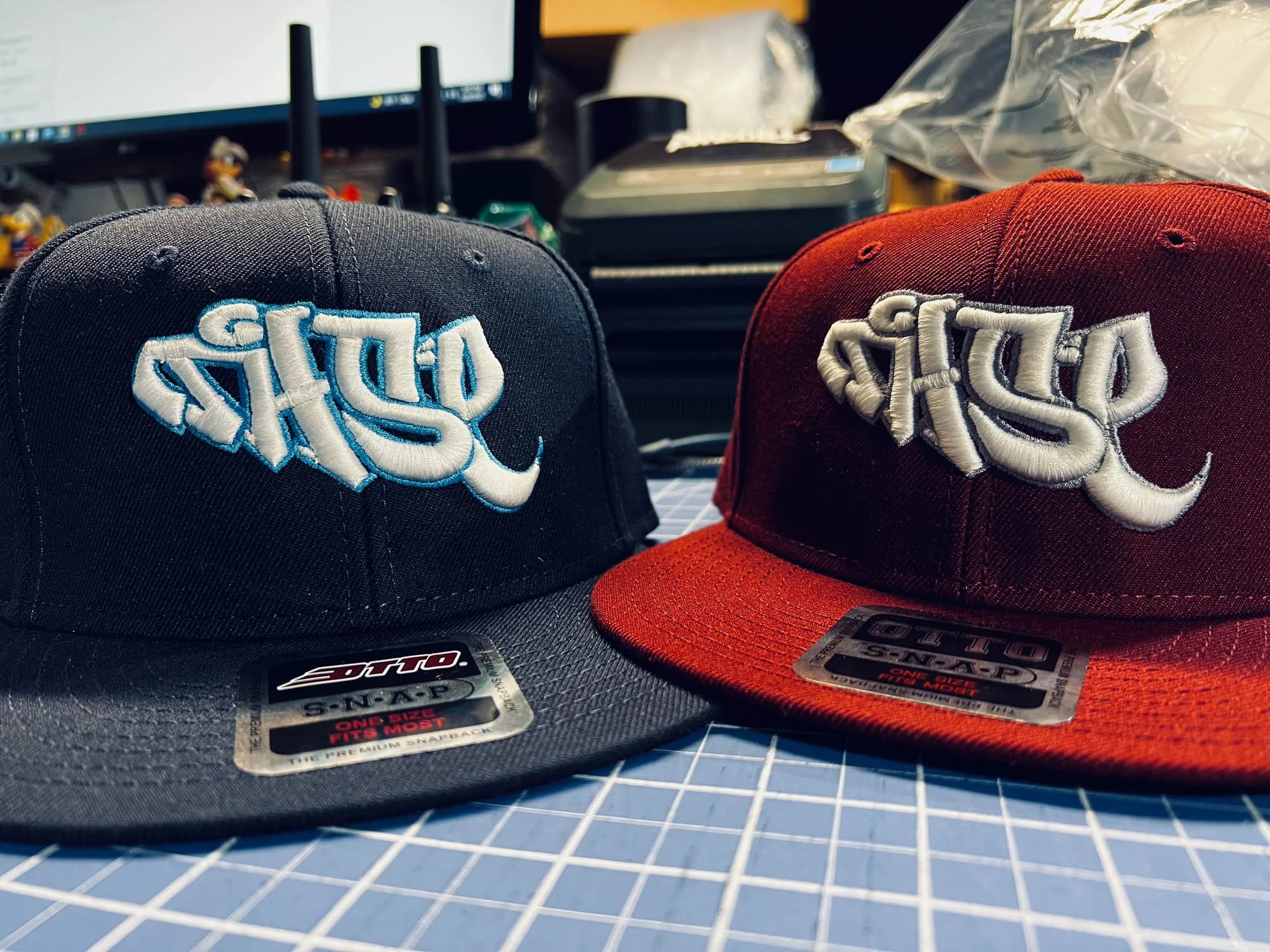 Two baseball caps, one black and one red, with white embroidered text, placed on a grid-lined surface, with a computer monitor and miscellaneous items in the background.