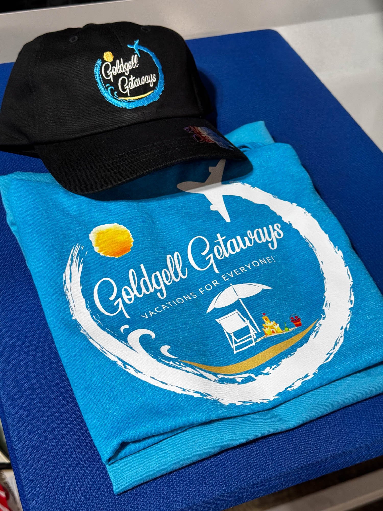 A black baseball cap with an embroidered logo reading 'Golddell Getaways' and a blue T-shirt with the same logo, a beach scene with an umbrella, sit on a blue surface.