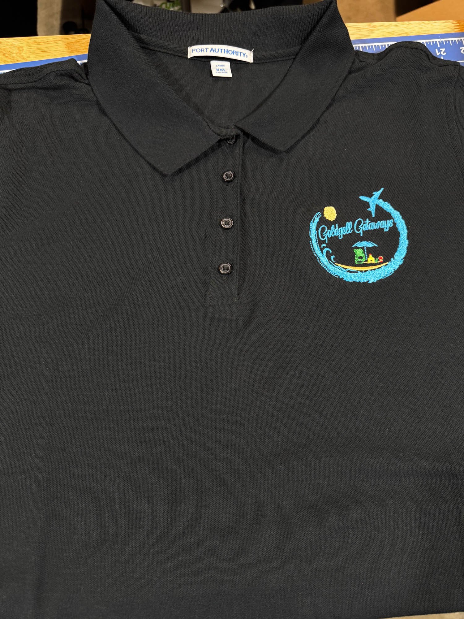 Black polo shirt with embroidered logo featuring a blue wave, a yellow sun, a green house, and a blue beach umbrella. The logo reads "Golddell Getaways."