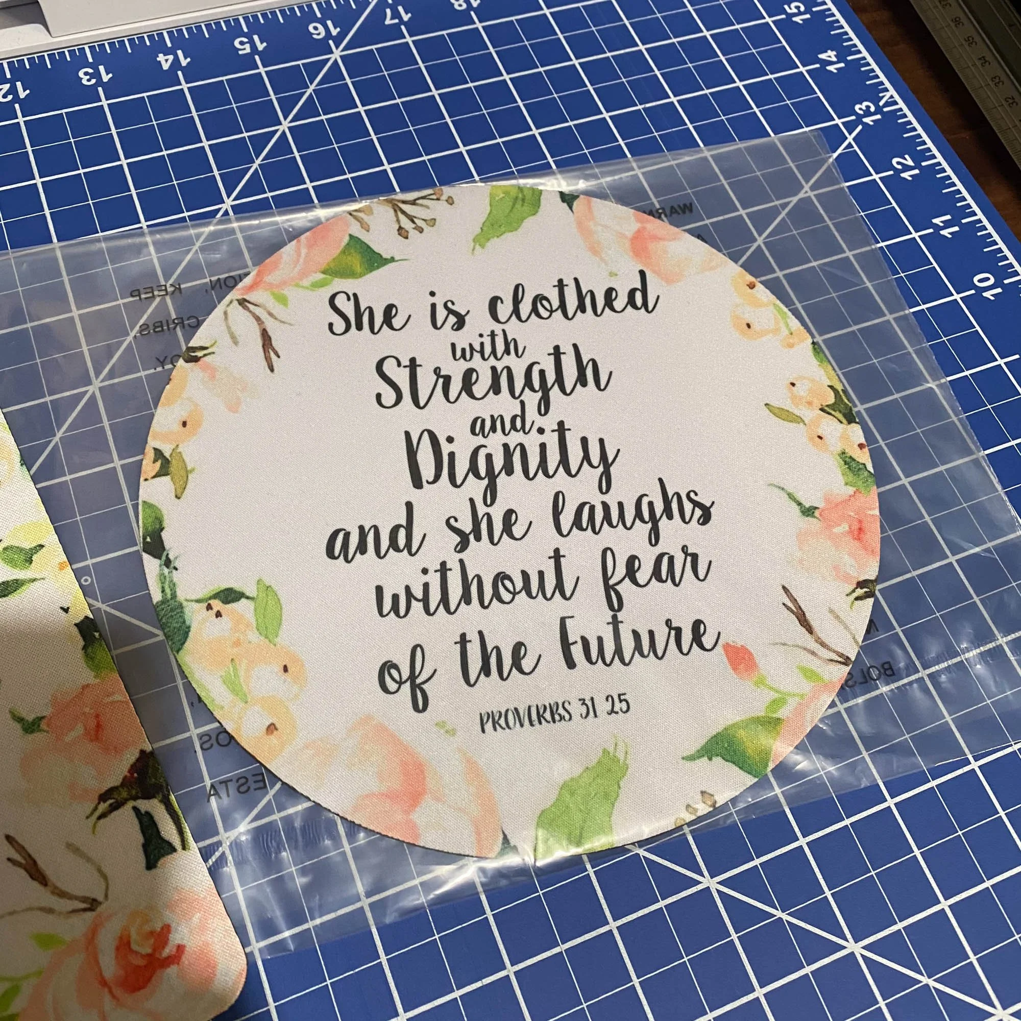 A round card with floral border on a grid cutting mat, displaying a quote from Proverbs 31:25 about strength, dignity, and laughter.