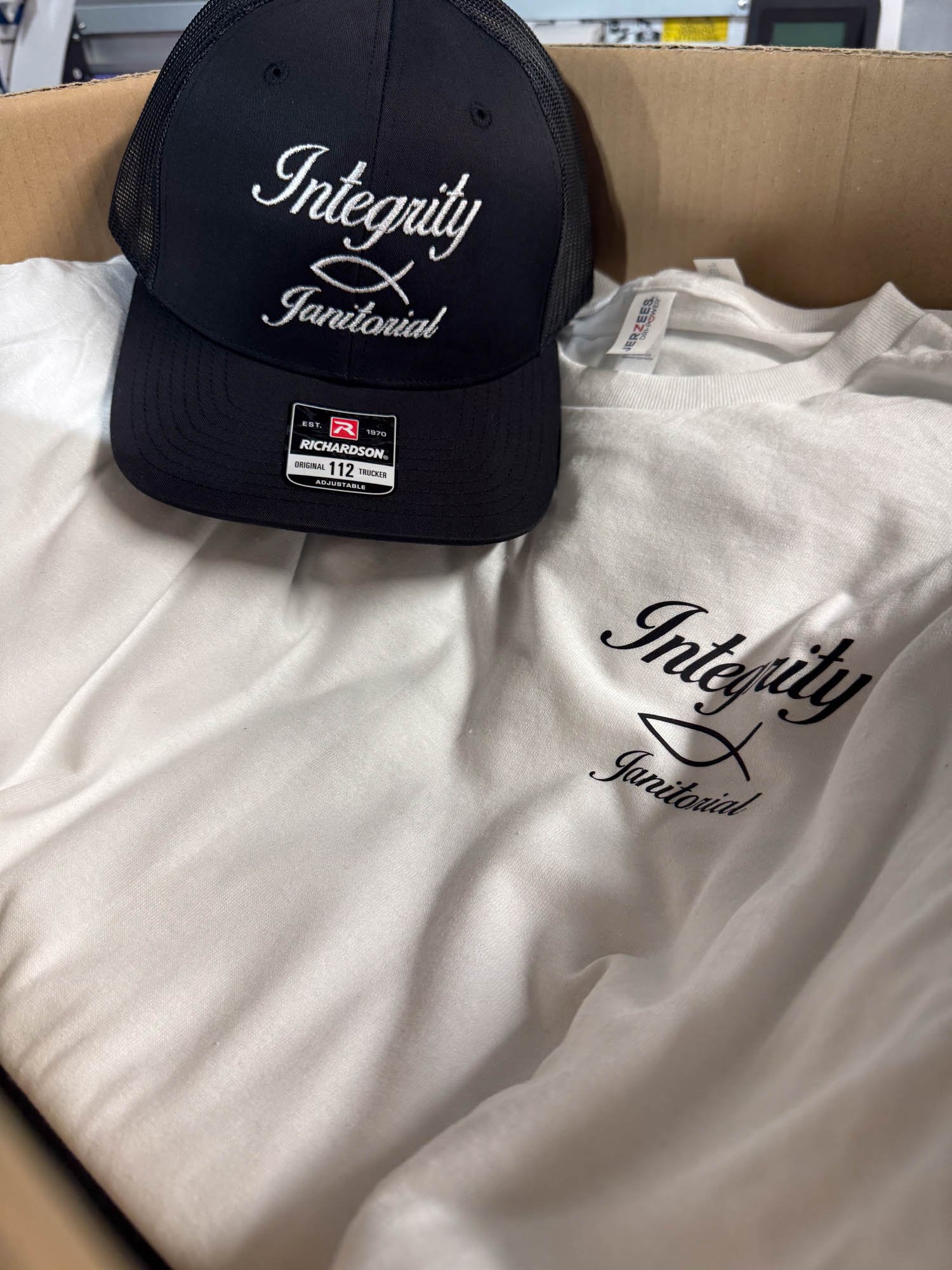 A black baseball cap with white embroidered text that says 'Integrity' and 'Tanitoutial', placed next to a white t-shirt with the same text in black, inside a cardboard box.