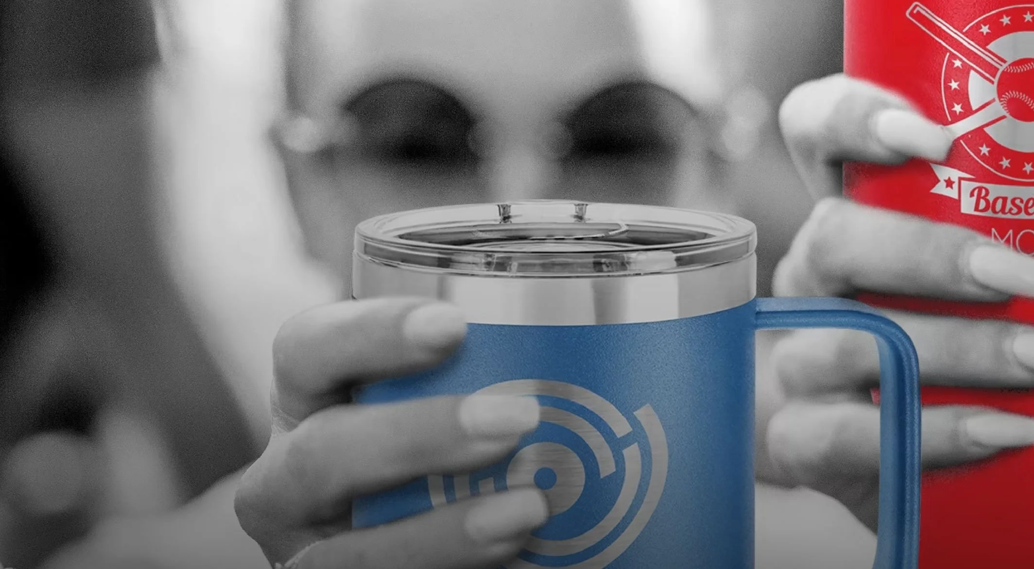 Close-up of two hands holding a blue mug and a red cup, with the background in black and white. The focus is on the colorful cups, which feature logos related to baseball, with the person's face blurred behind them.