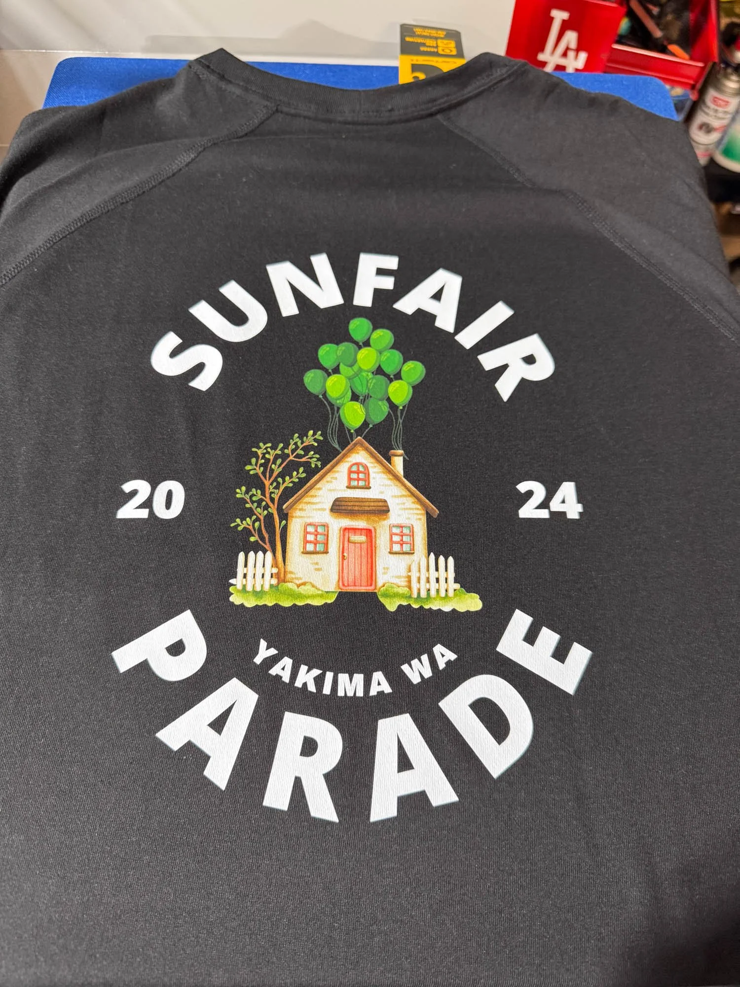 Black t-shirt with a graphic design of a house with a tree and balloons, and text 'SUNFAIR 2024 PARRAD YAKIMA WA' arranged in a circular pattern.